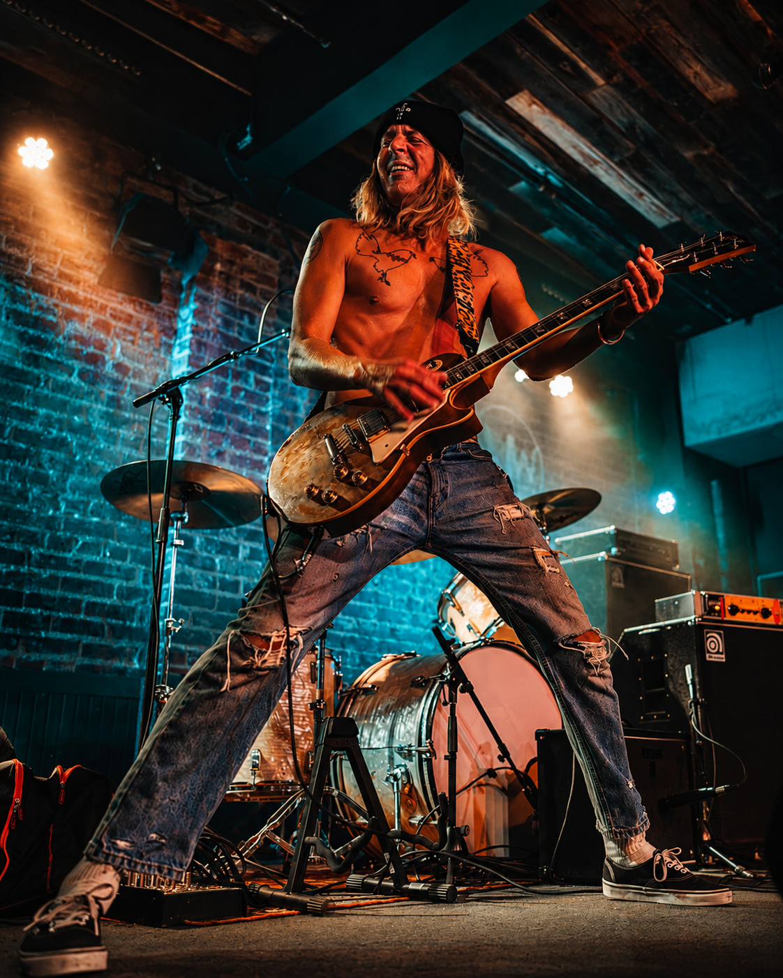 Shirtless male guitarist performing on stage with electric guitar, dramatic lighting, brick wall background.