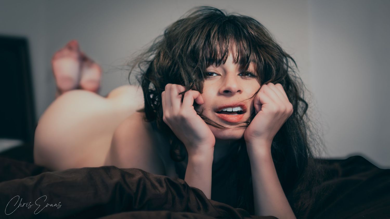 A person lies on dark sheets while smiling and resting their chin on their hands in a boudoir style portrait.