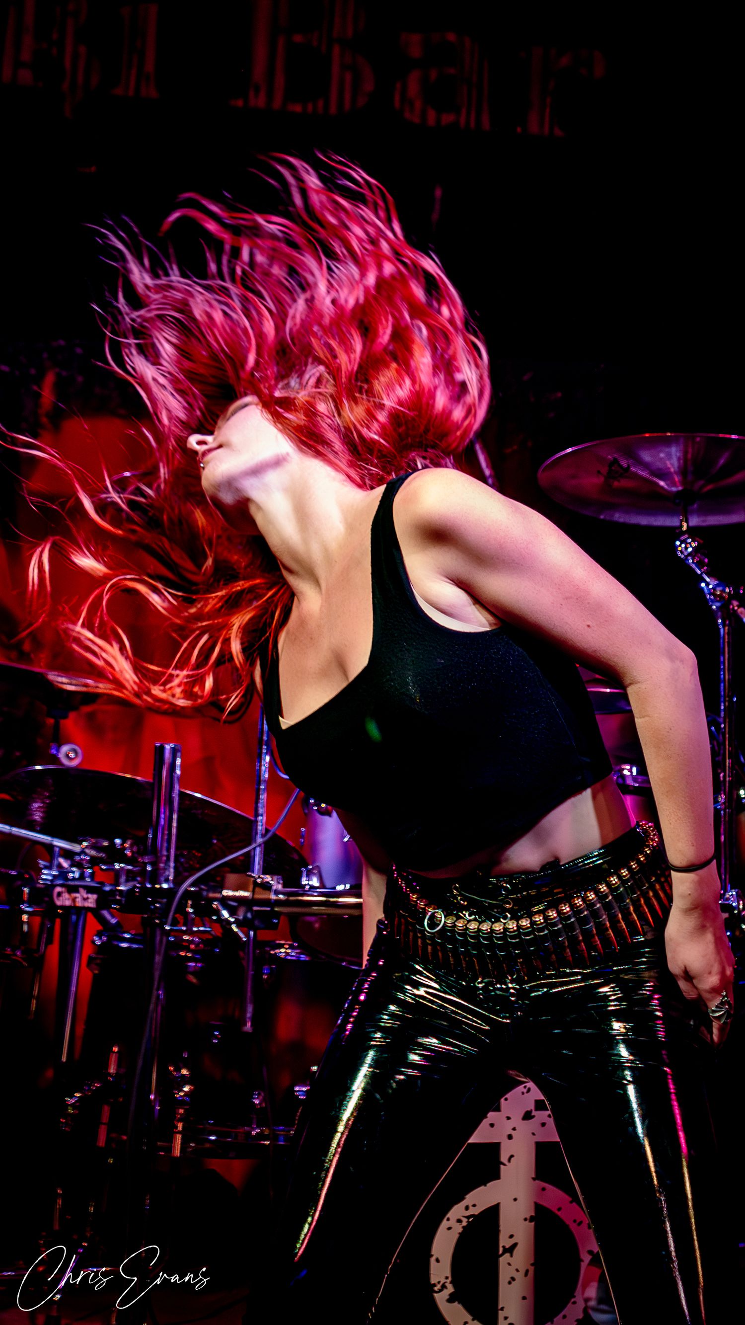 A performer with vibrant red hair flips their head back on stage, wearing black clothing with a drum kit visible in background.