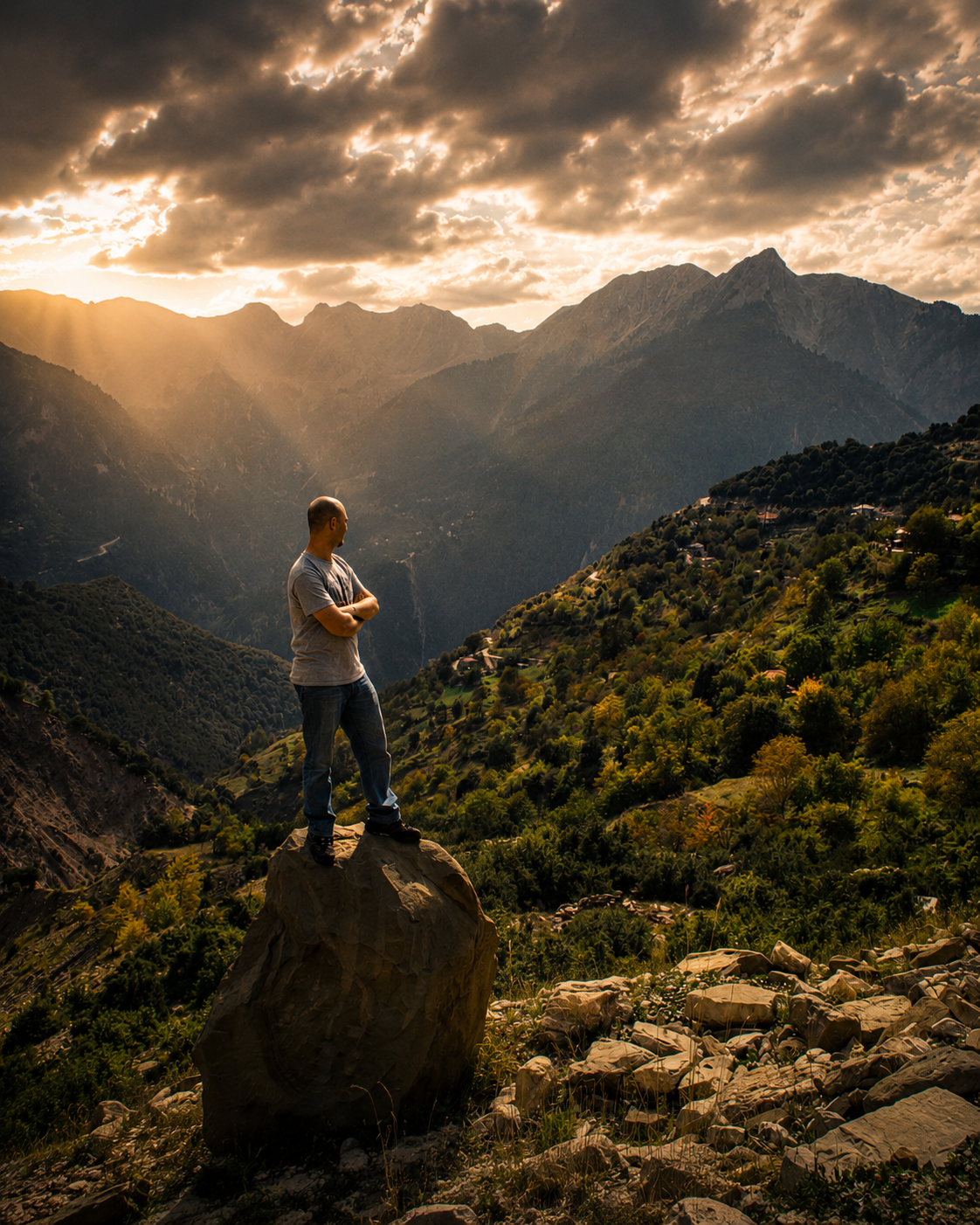 A lone hiker stands on a boulder overlooking a dramatic mountain valley at golden sunset with rays piercing stormy clouds.