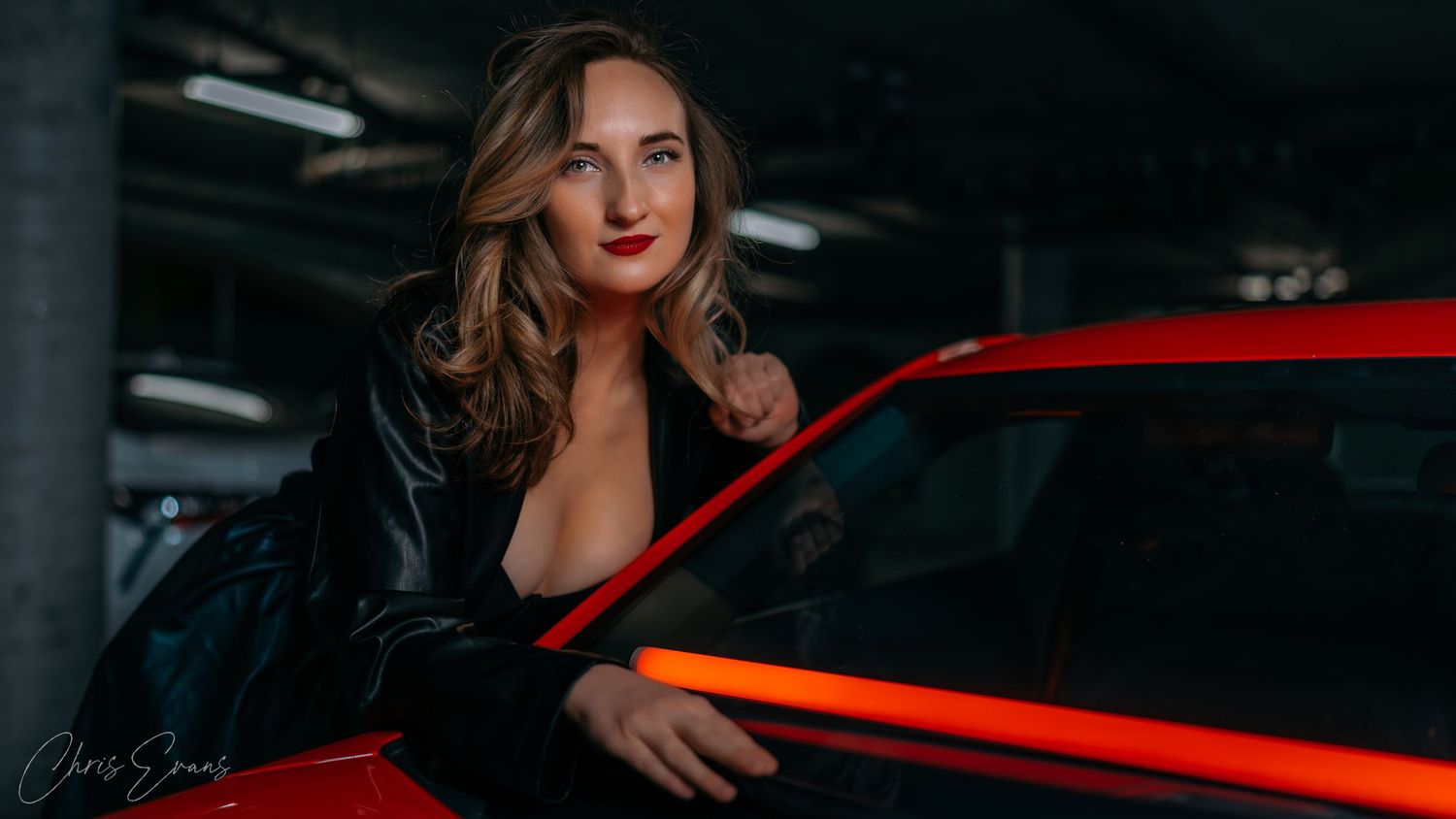 Dramatic portrait of woman in black jacket posed next to red sports car in dark parking garage.