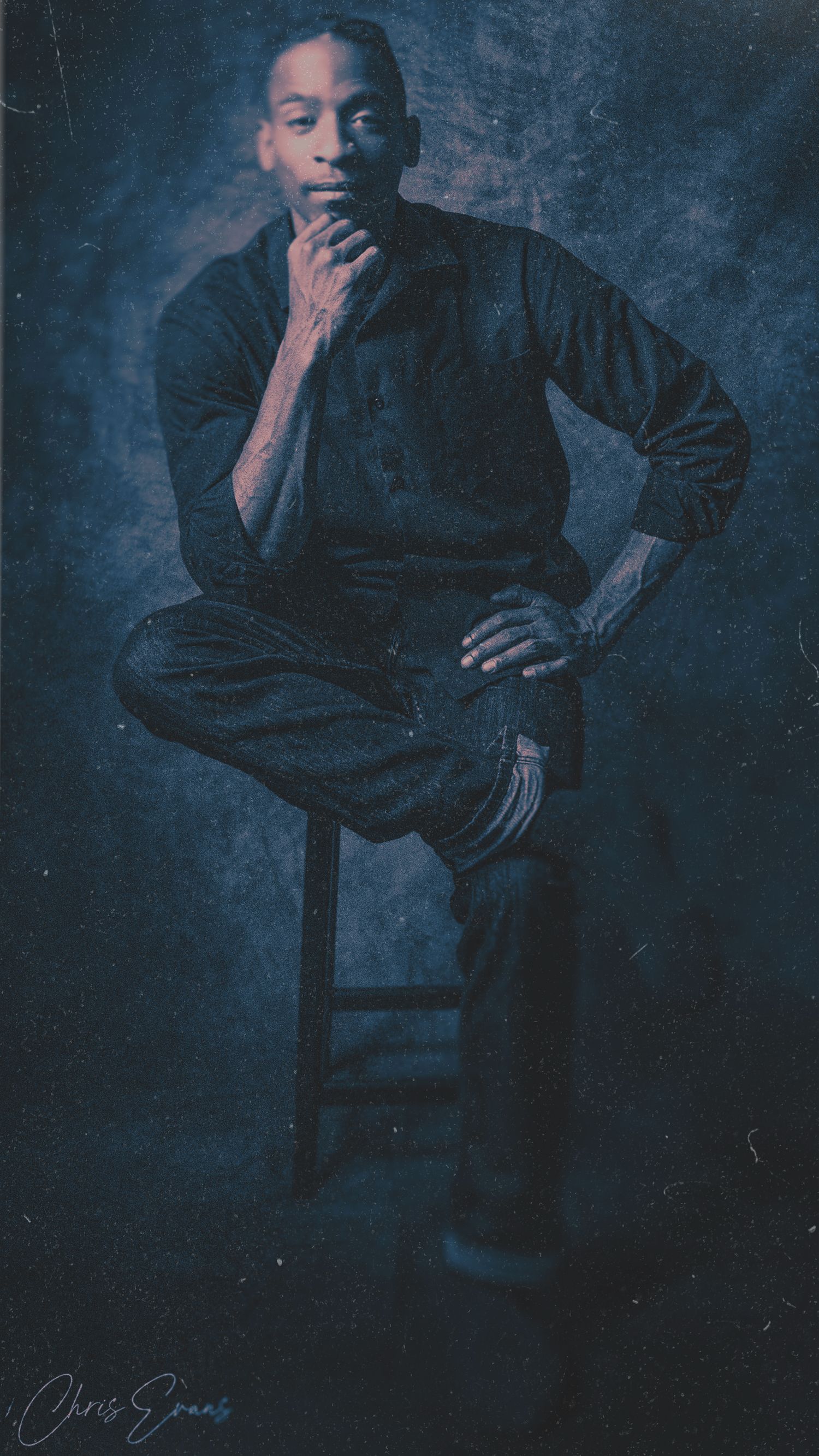 Artistic portrait in moody blue tones showing a contemplative figure seated on a stool against a dark background.