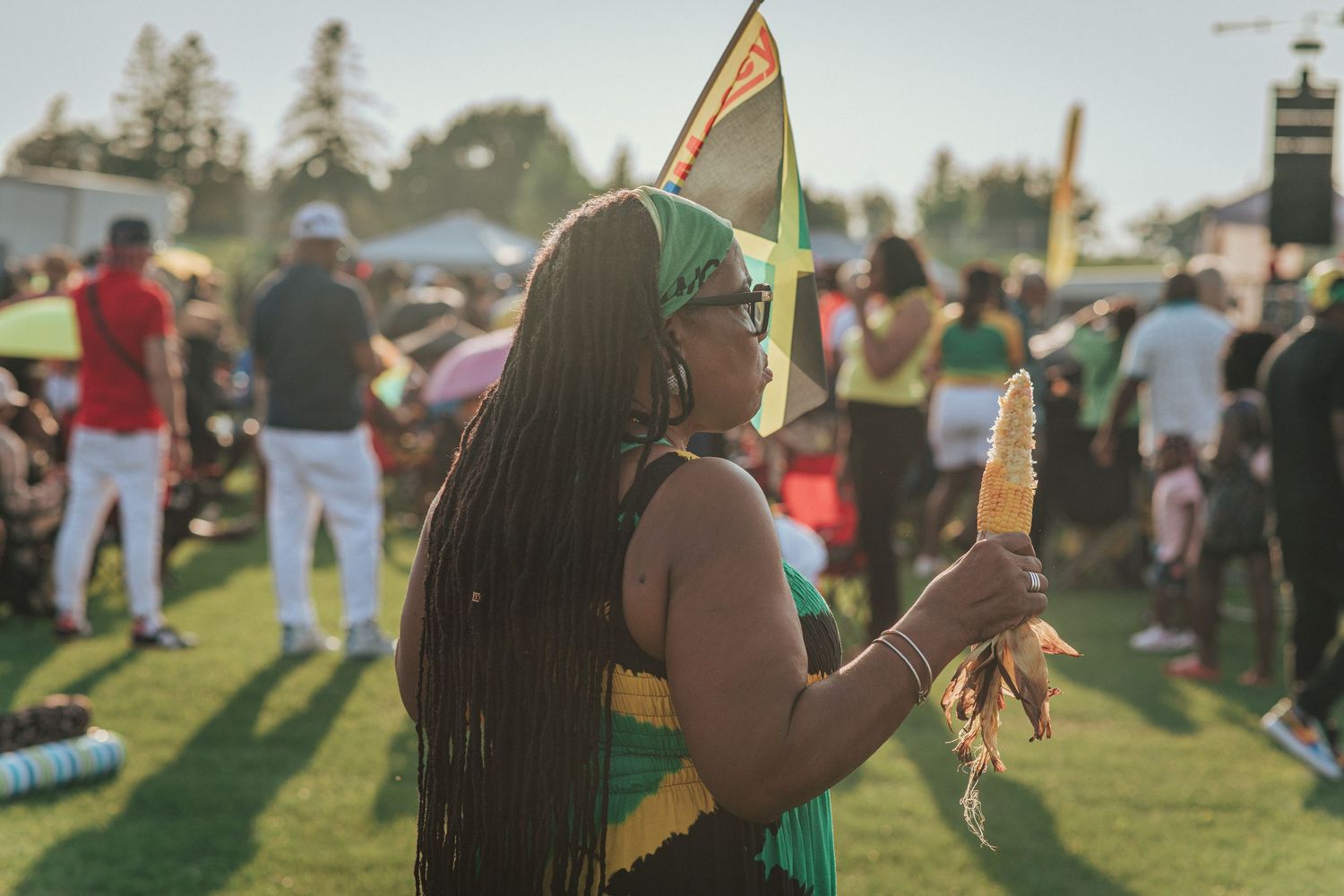 A black woman wearing a Jamaica-themed crochet dress, holding a roasted corn while waving a Jamaica flag at the Jamaica Day Toronto Festival