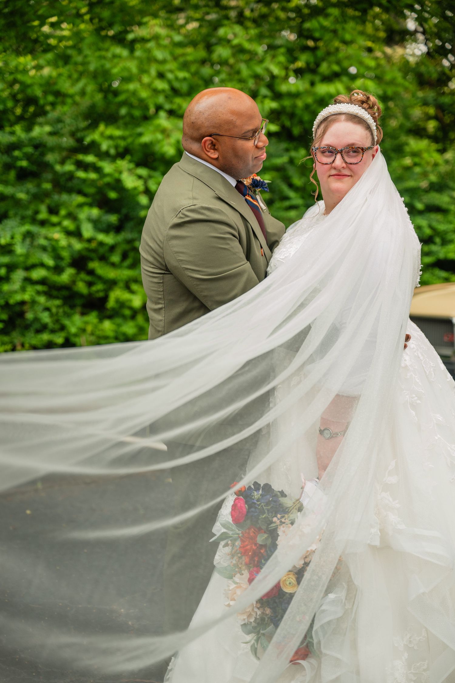 Flowing white wedding dress veil billowing dramatically in the wind against lush green foliage background.