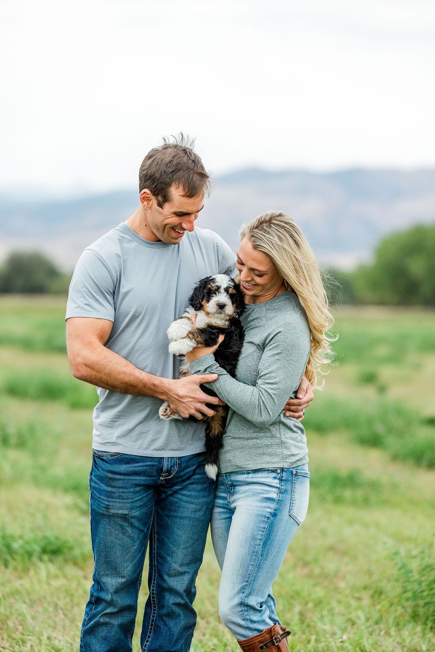 Couple in matching blue outfits embrace while holding their small black and white dog in a green field.