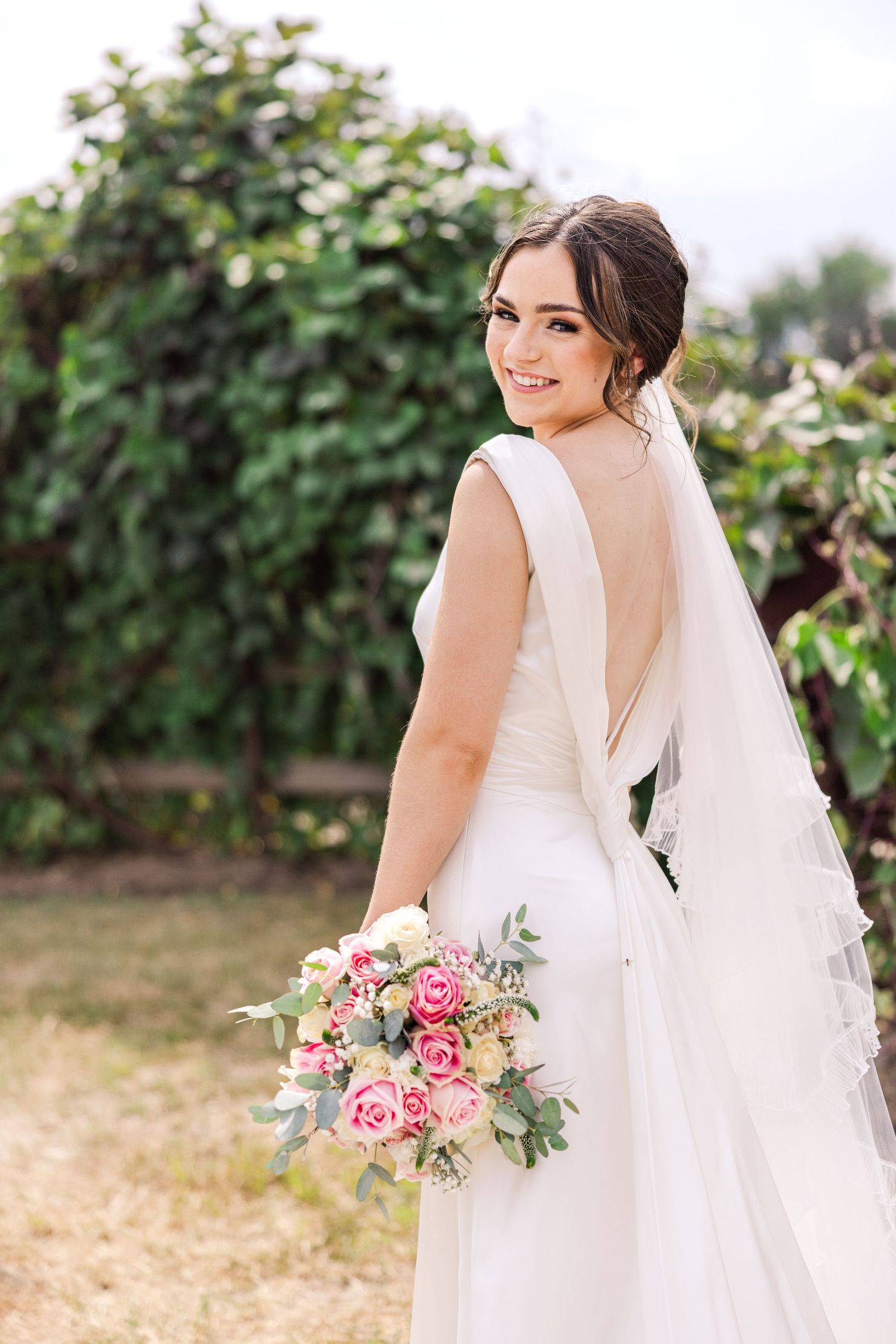 Bride in white backless gown holding pink rose bouquet, smiling over shoulder in lush garden setting.