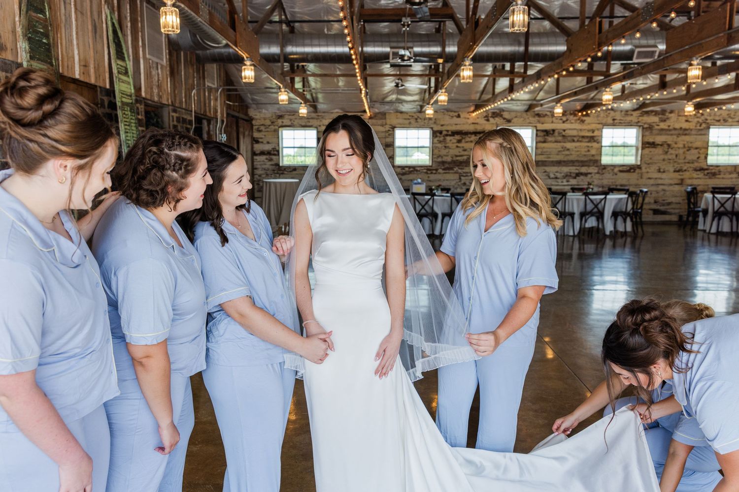 Bride in white gown with veil surrounded by bridesmaids in light blue dresses inside a rustic barn venue.