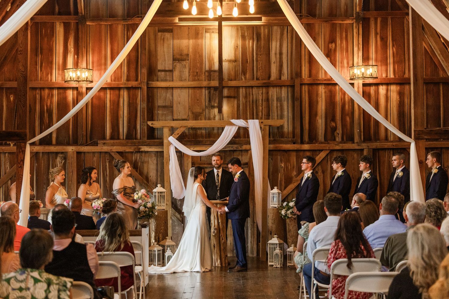 Rustic barn wedding ceremony with couple at floral arch altar, chandelier lighting, wood walls, and seated guests.