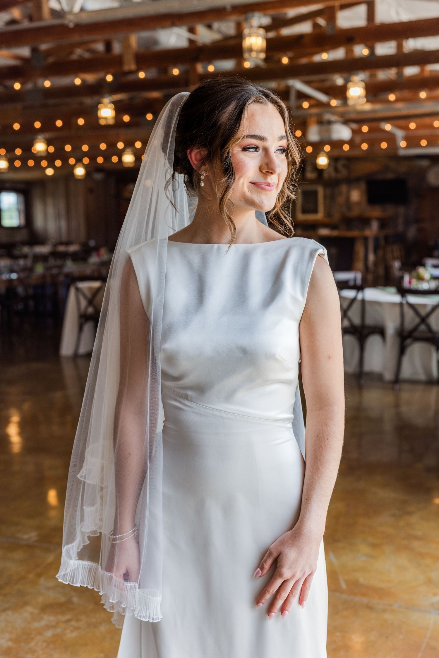 Beautiful bride in minimalist satin wedding dress and veil poses in warmly lit rustic barn venue.