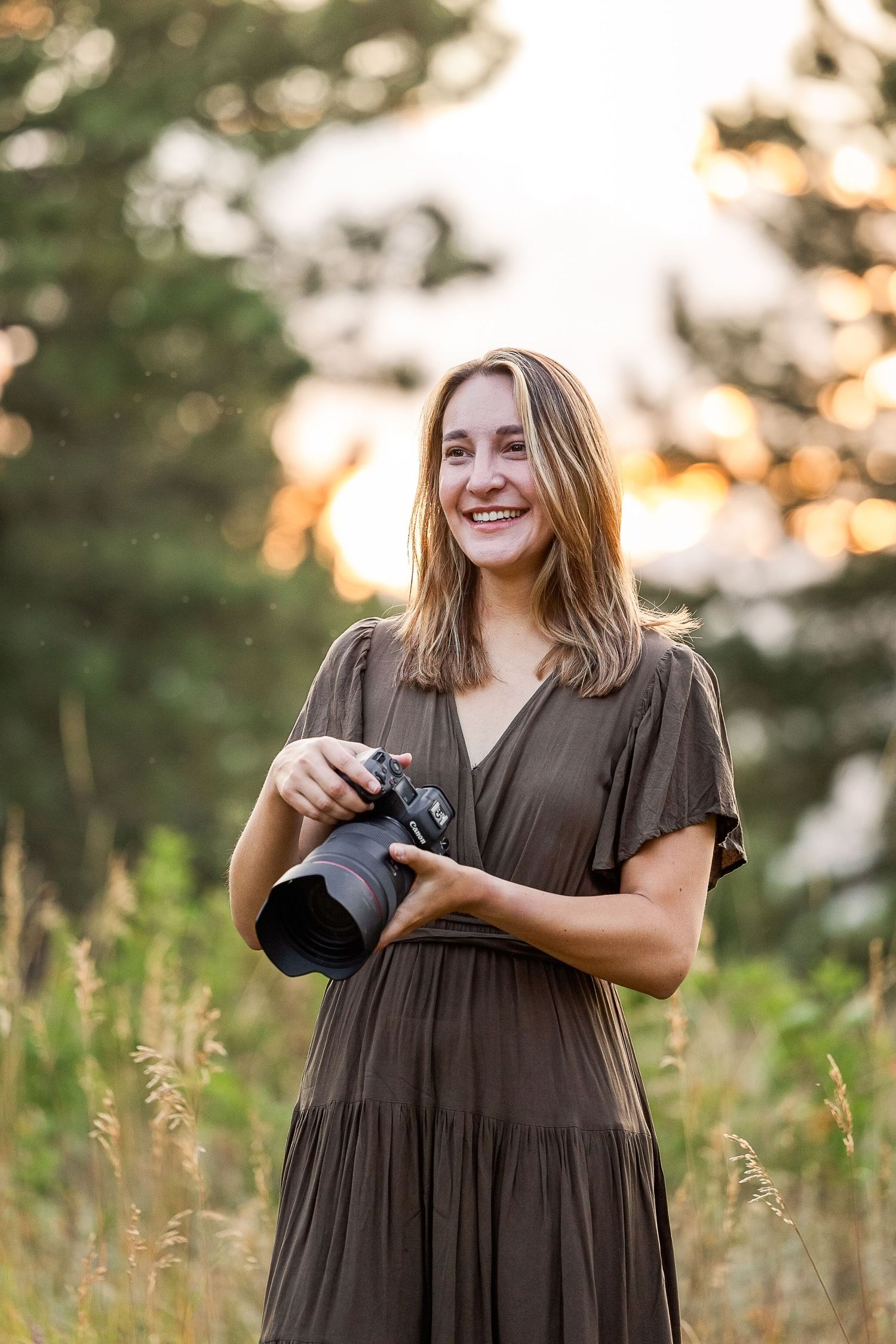 A photographer in a brown dress stands in a golden field at sunset holding a camera.