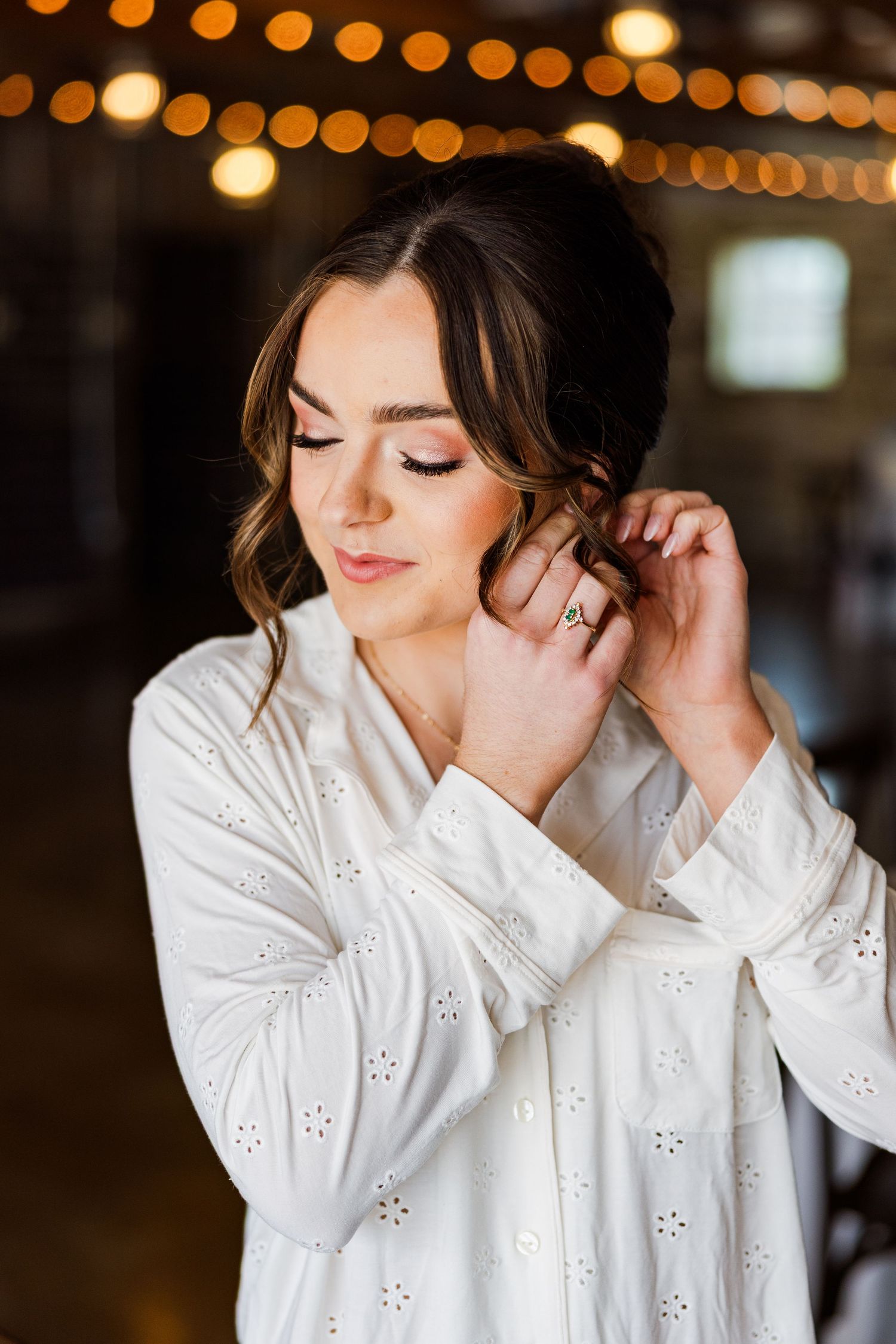 Bride in white eyelet dress adjusting earring with soft glam makeup and updo hairstyle, bokeh lights in background.