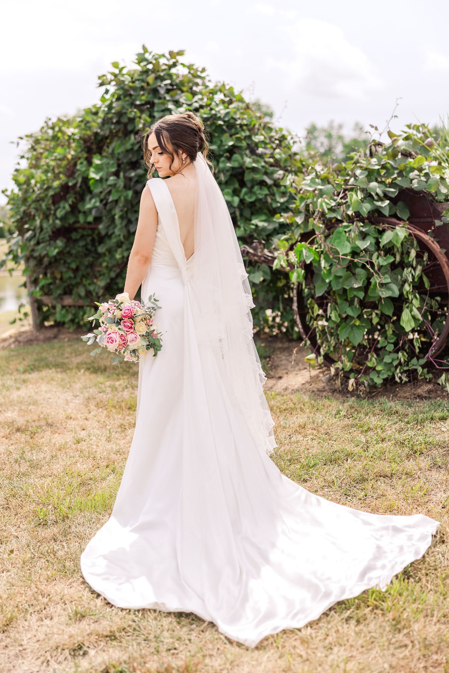 Bride in elegant white gown with open back and veil holds bouquet outdoors near lush green vineyard.
