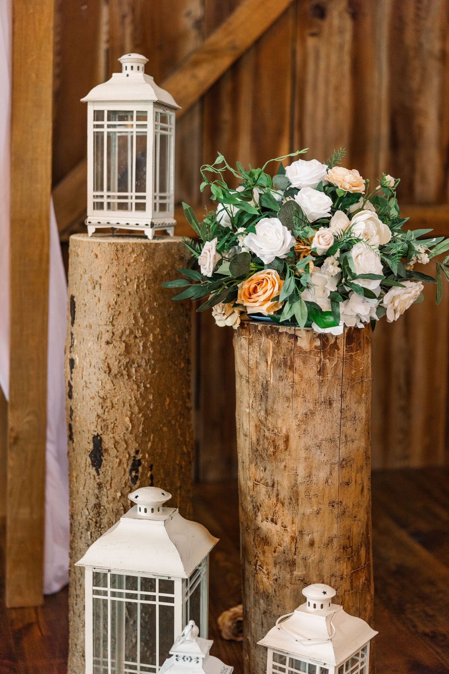 Rustic wedding decor with white lanterns on wood floor and floral arrangement of white and peach roses on log pedestals.