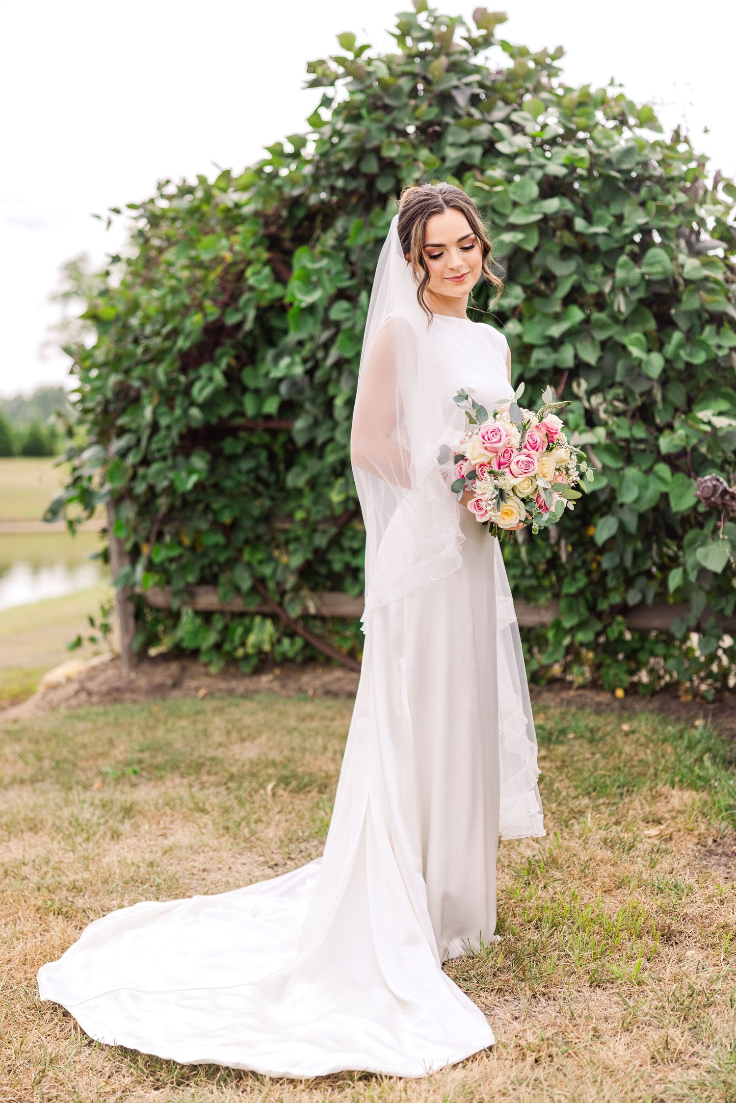 Bride in elegant white gown with long train and veil holds pink bouquet outdoors near lush green arch.