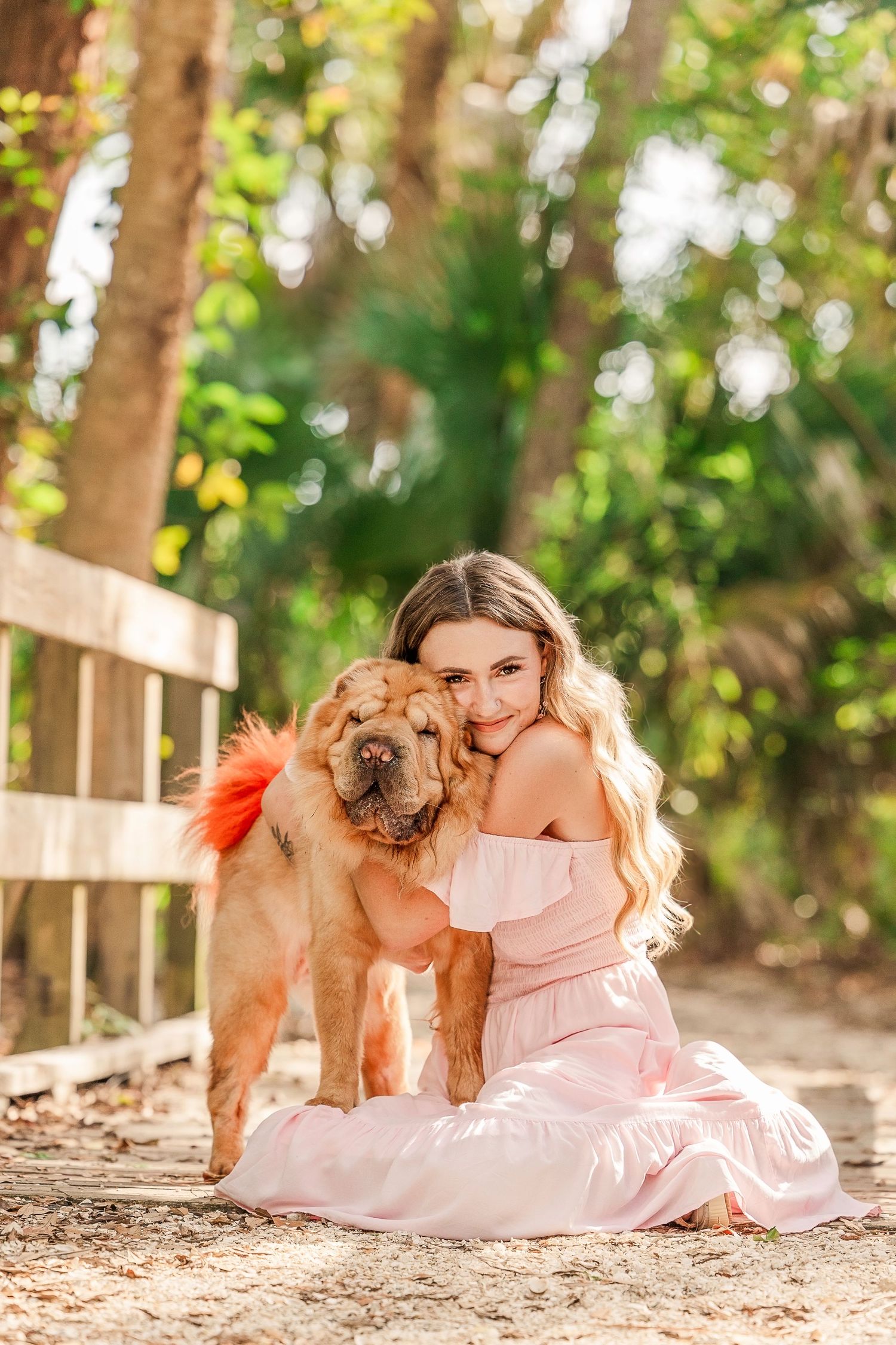 Young woman in pink off-shoulder dress hugging a fluffy Chow Chow puppy outdoors in a sunny park setting.