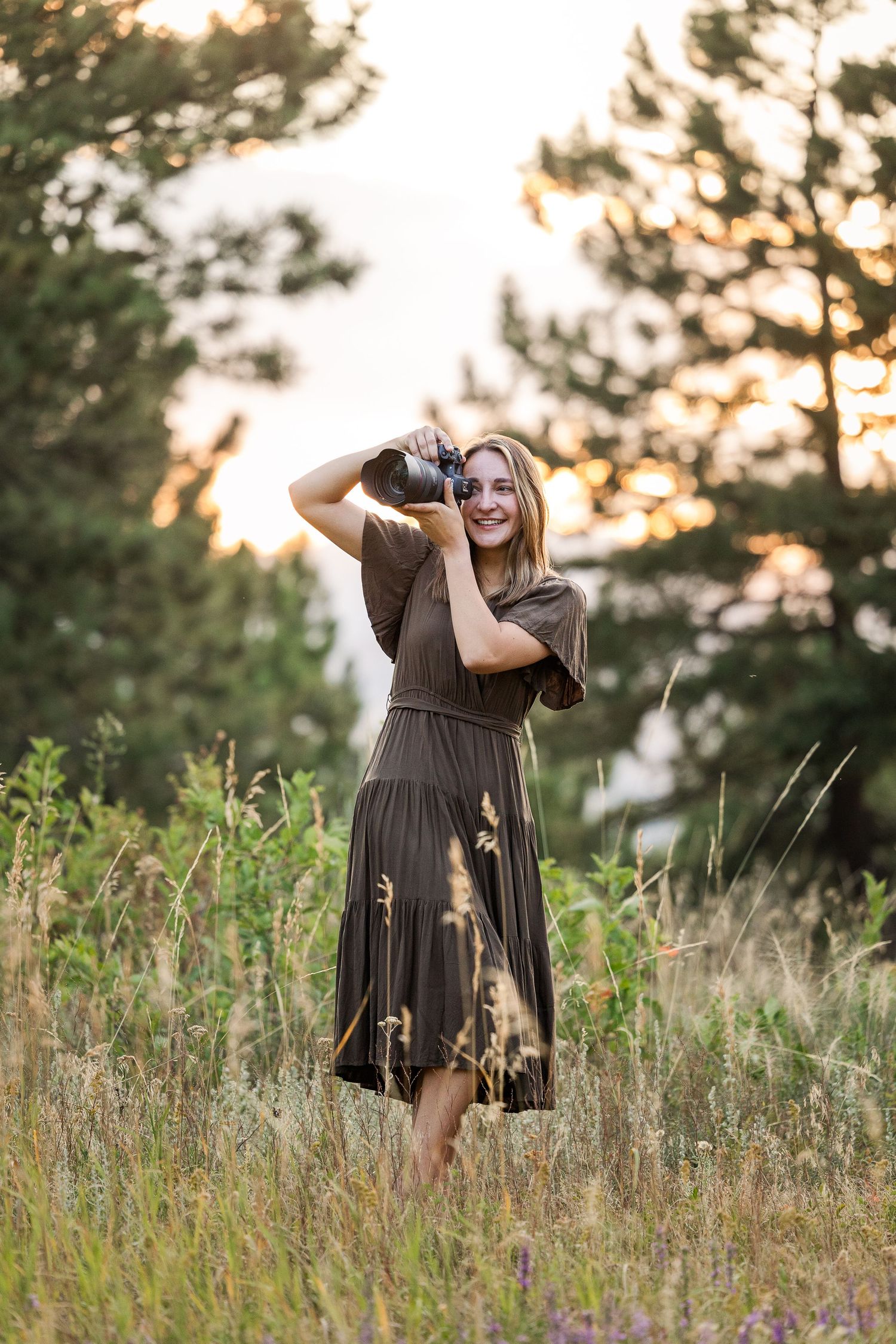Someone in a brown dress poses playfully in a wildflower meadow at sunset with tall pine trees in the background.
