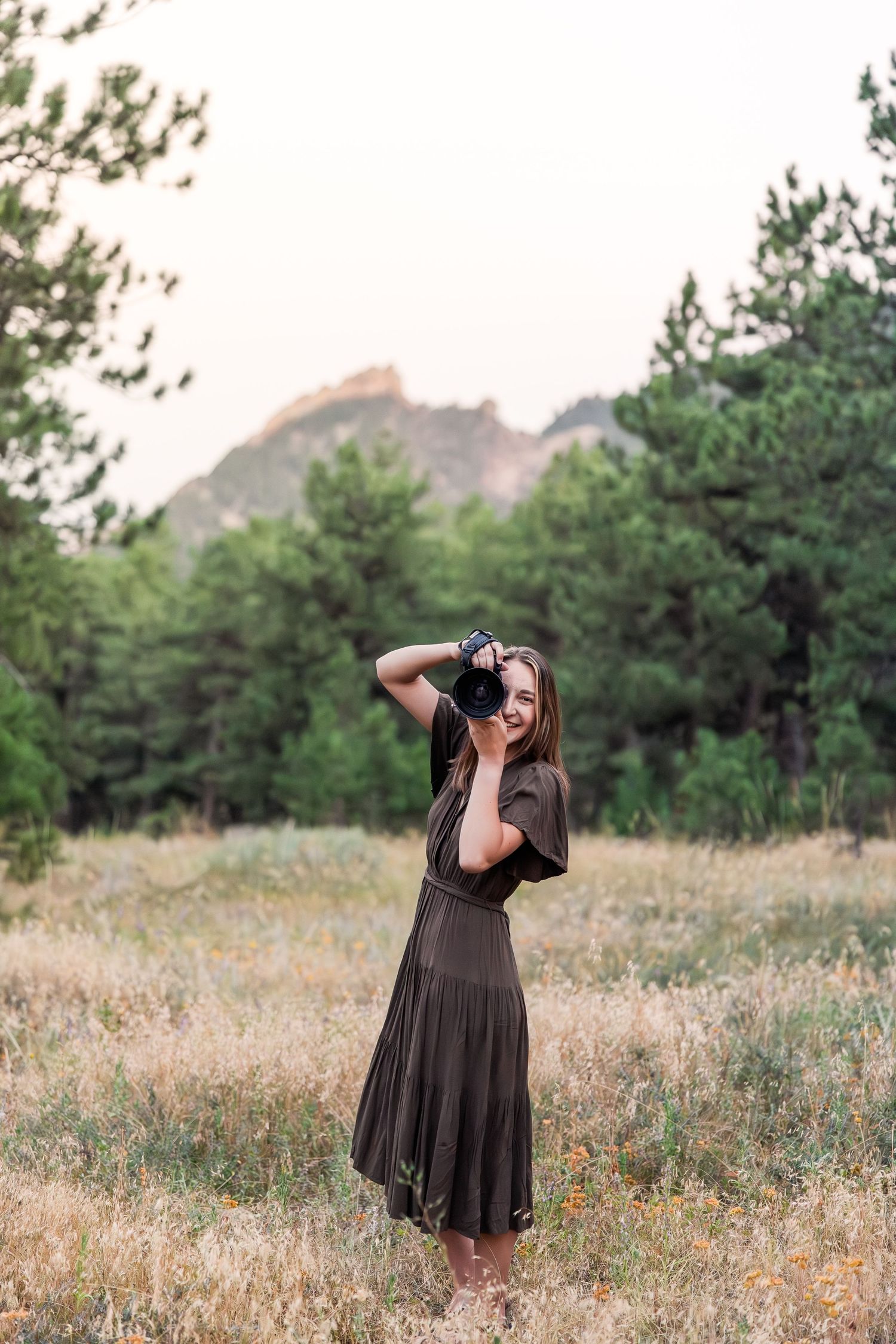 A person in a flowing black dress twirls gracefully in a wildflower meadow with pine trees and mountains in the background.