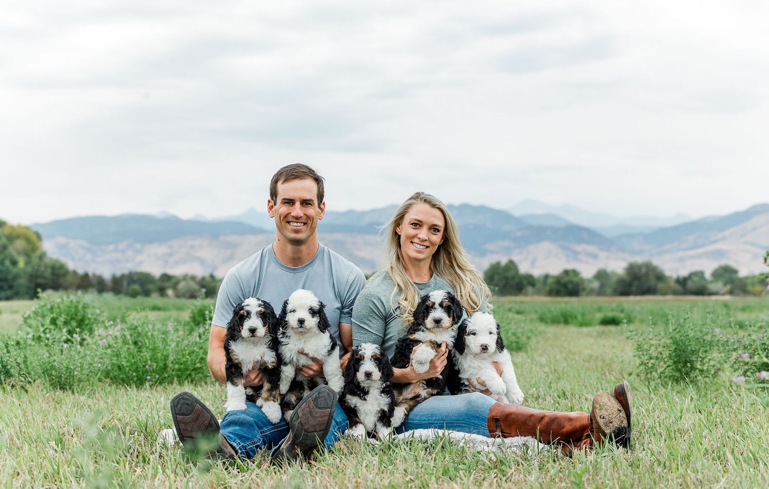 A couple sits on a blanket in a grassy field with their two Husky dogs against a scenic mountain backdrop.