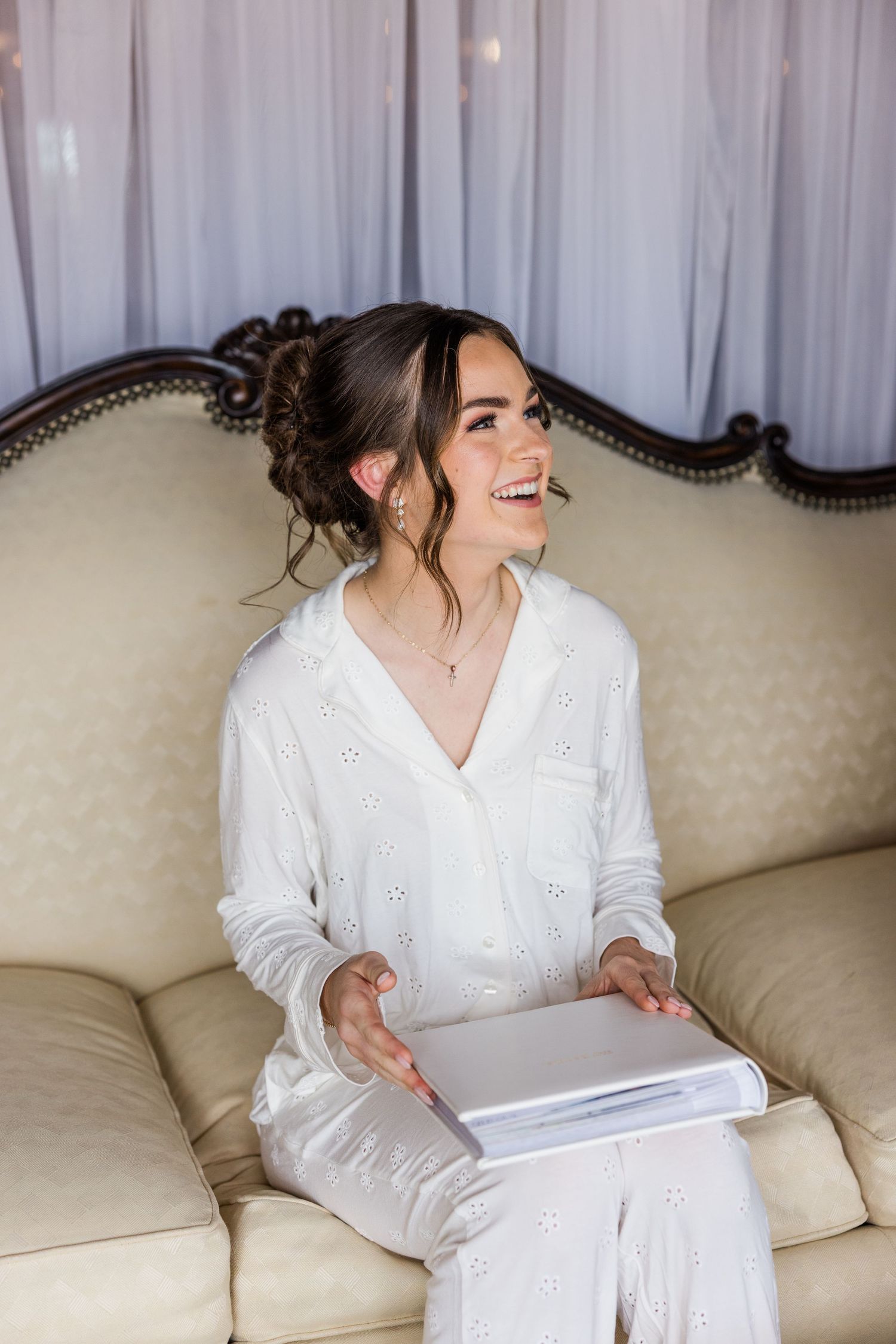 Joyful bride in white eyelet pajamas laughs while holding wedding album on cream vintage sofa.