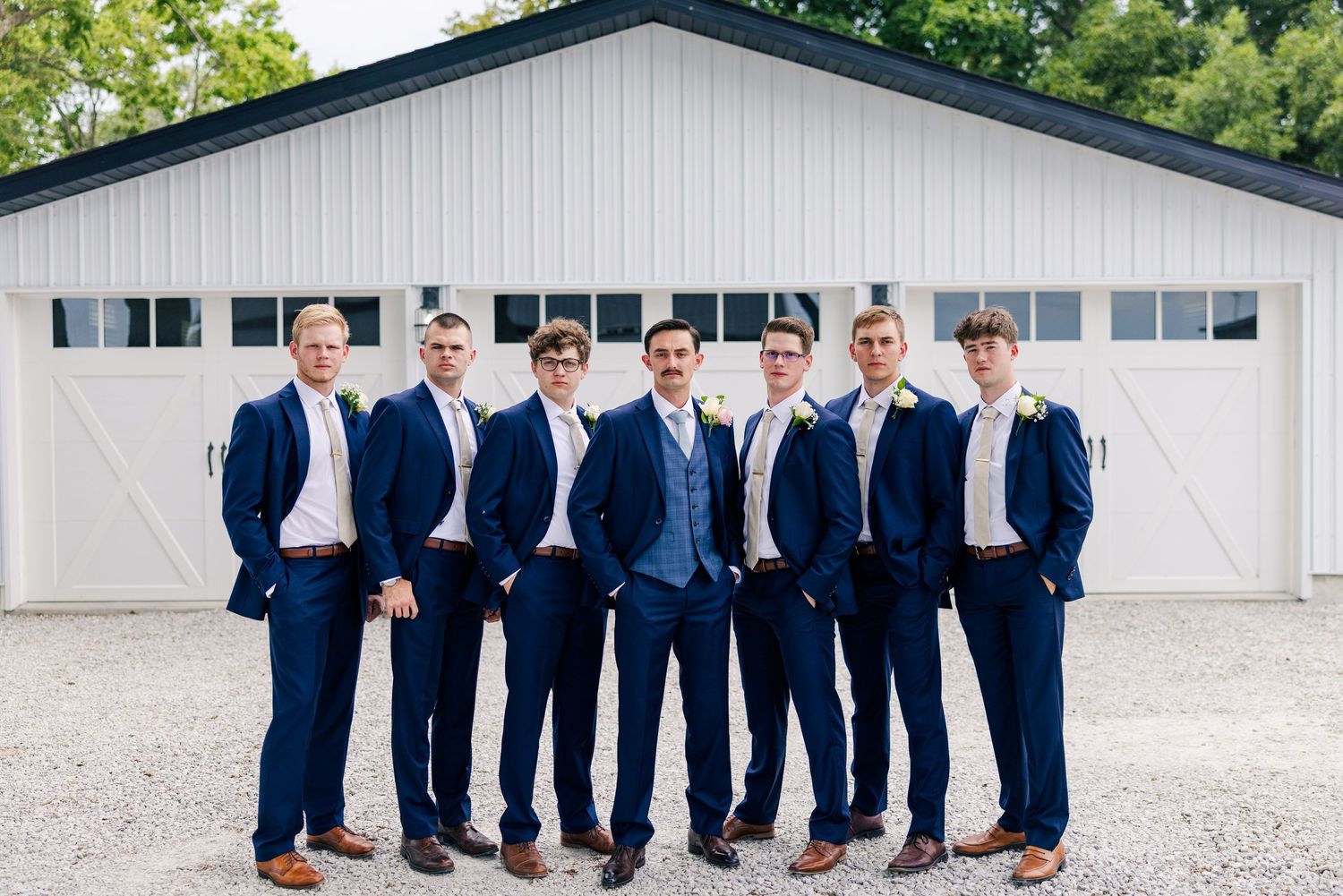 Seven groomsmen in navy blue suits with boutonnieres pose in front of a white barn at an outdoor wedding.