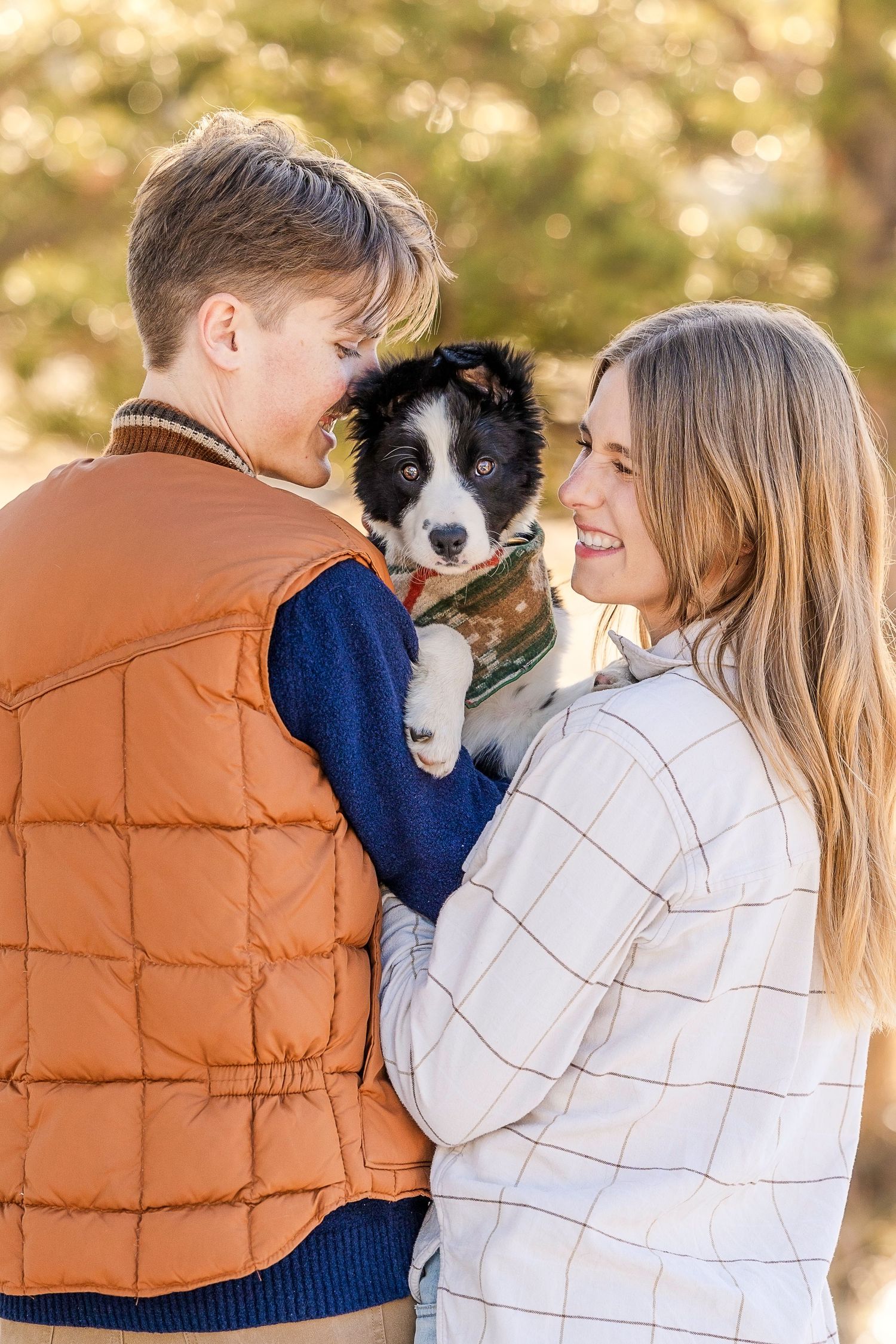 A young couple smiling at each other while holding a black and white border collie puppy outdoors in autumn.