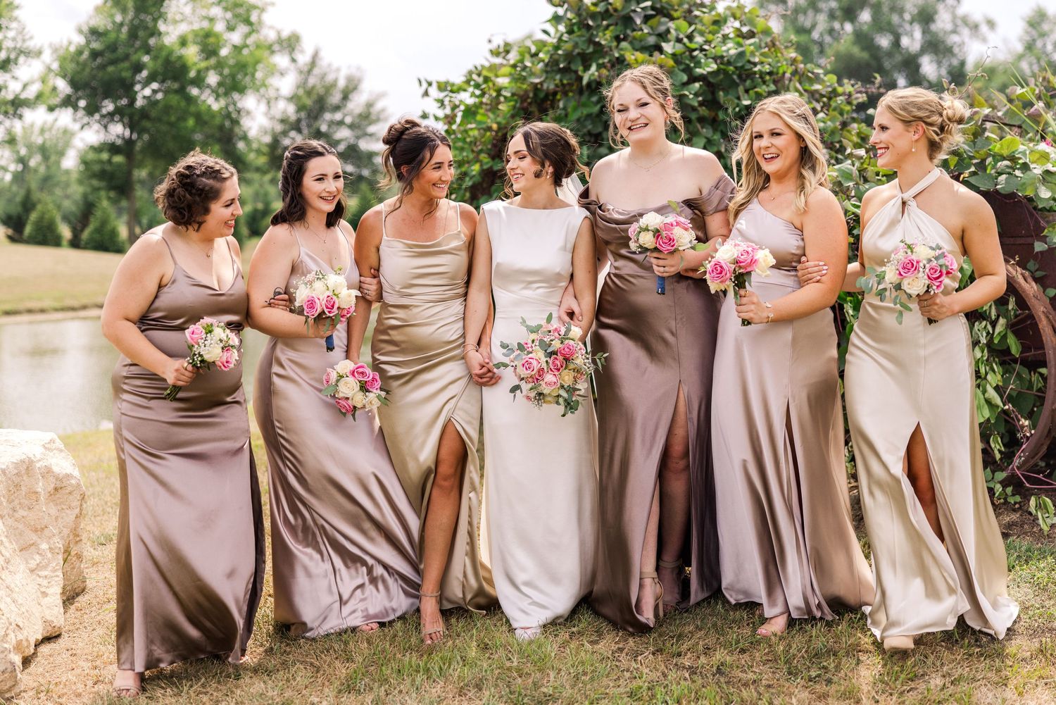 Bride and six bridesmaids in champagne satin gowns laugh together outdoors, holding pink and white bouquets.