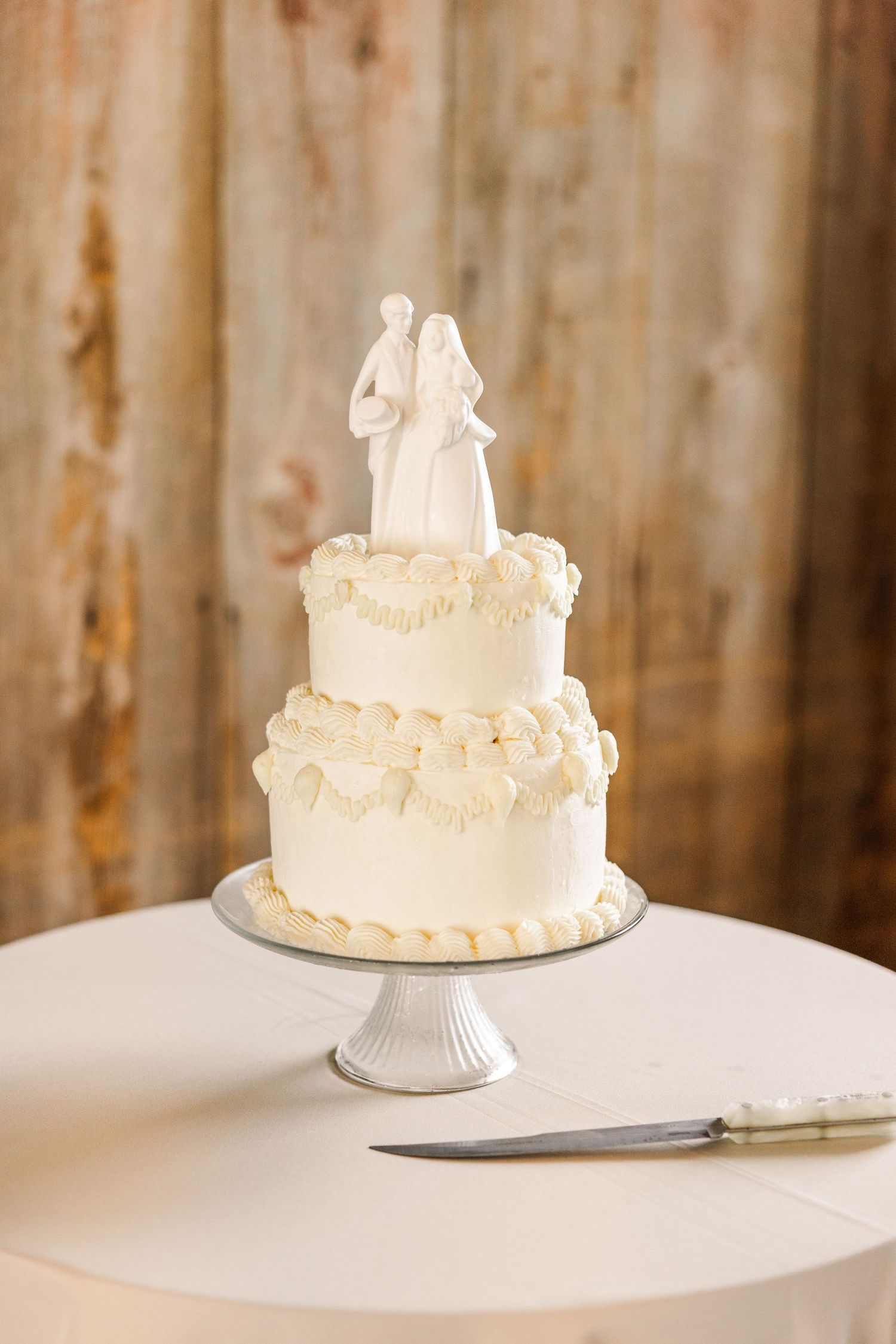 Elegant two-tier white wedding cake with bride and groom topper and ruffled frosting on a silver cake stand.