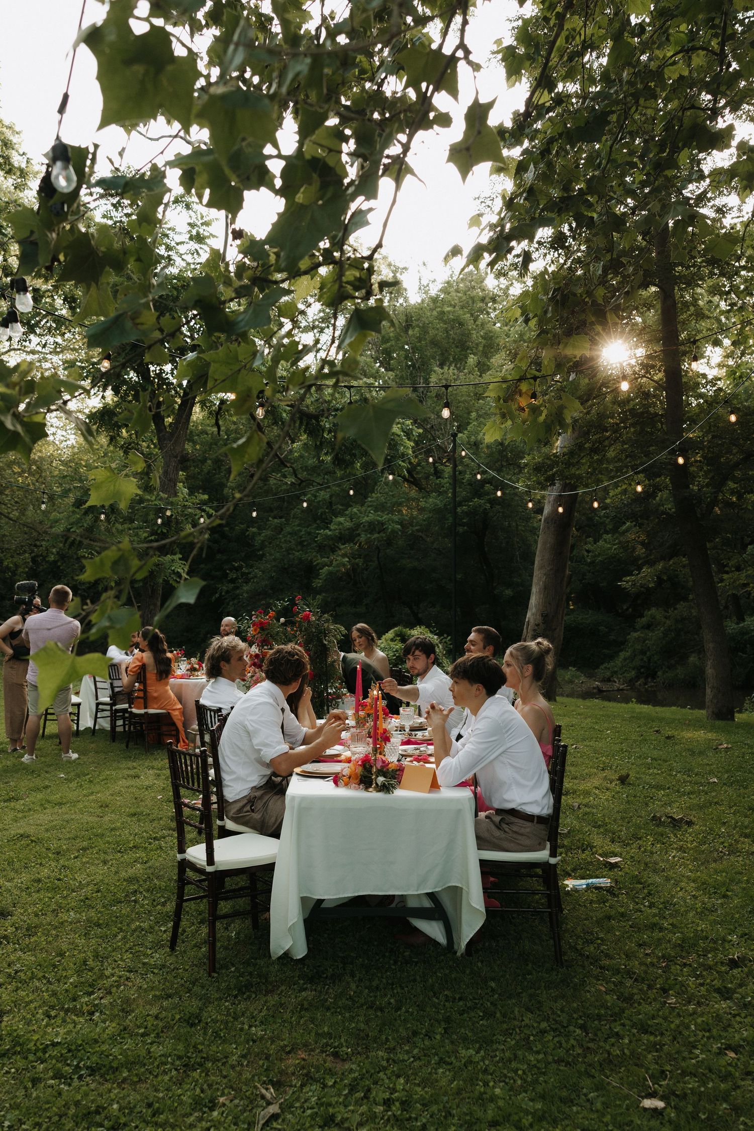 Outdoor dinner party setup with long tables and chairs arranged under trees at sunset in a garden setting.