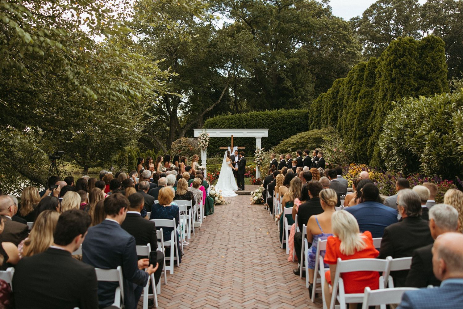 Guests seated in rows at an outdoor garden wedding ceremony with lush greenery and trees in the background.