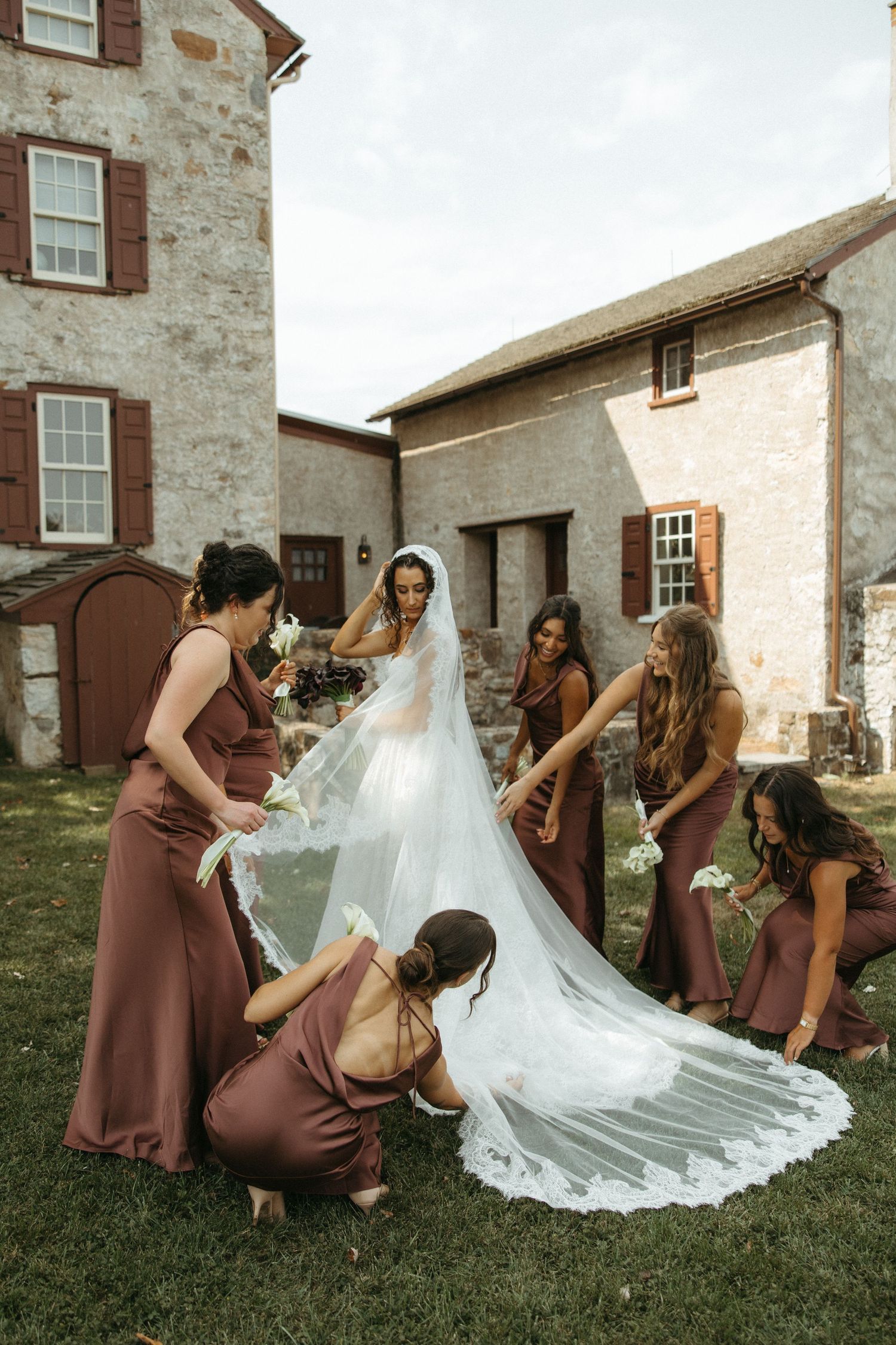 Bridesmaids in burgundy dresses help arrange a bride's long veil on grass outside a stone building.