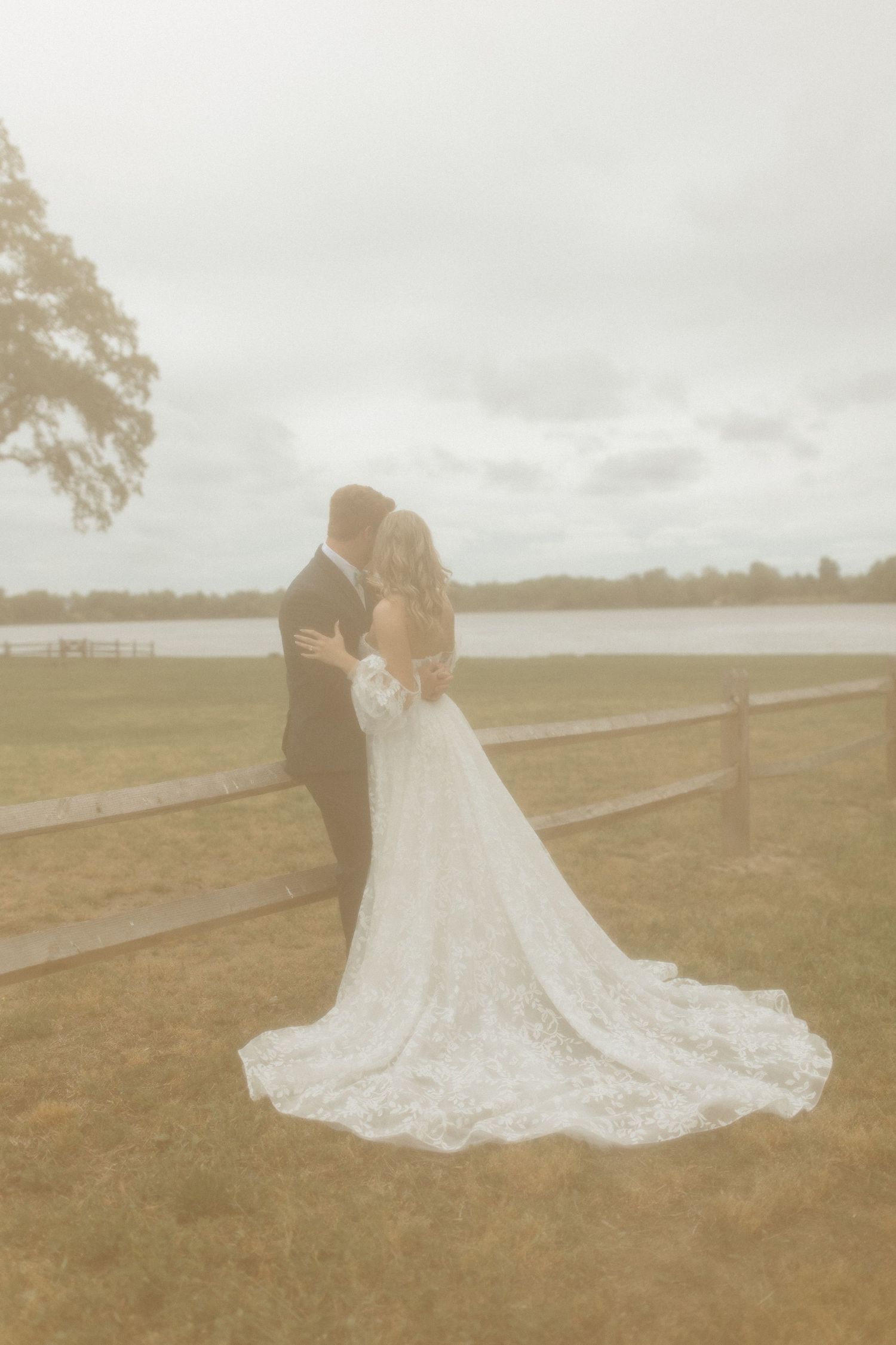 A couple in wedding attire embraces on a grassy field with a lake and trees in the background.