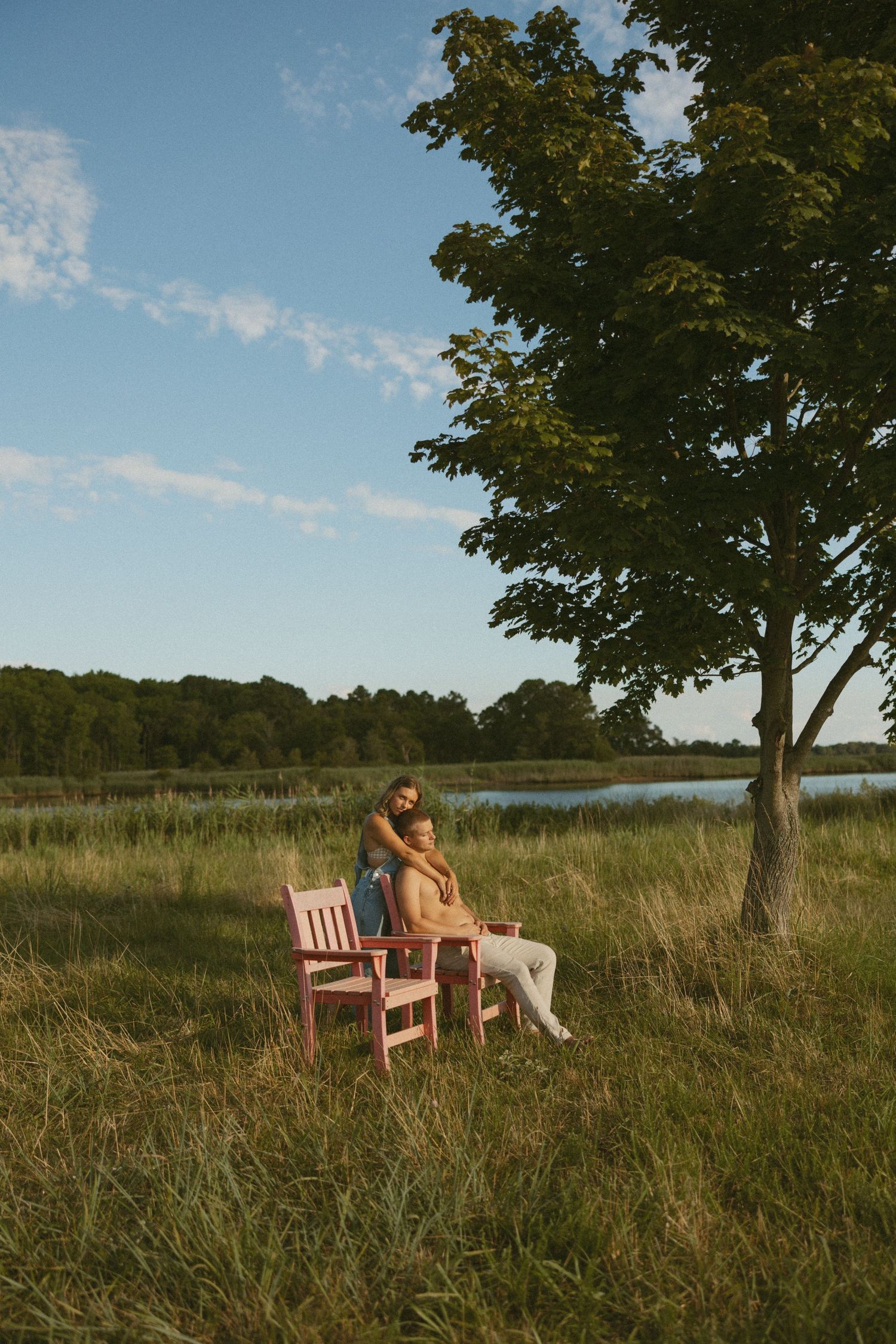 Couple relaxing on wooden bench in tall grass meadow by lake during golden hour with trees in background.