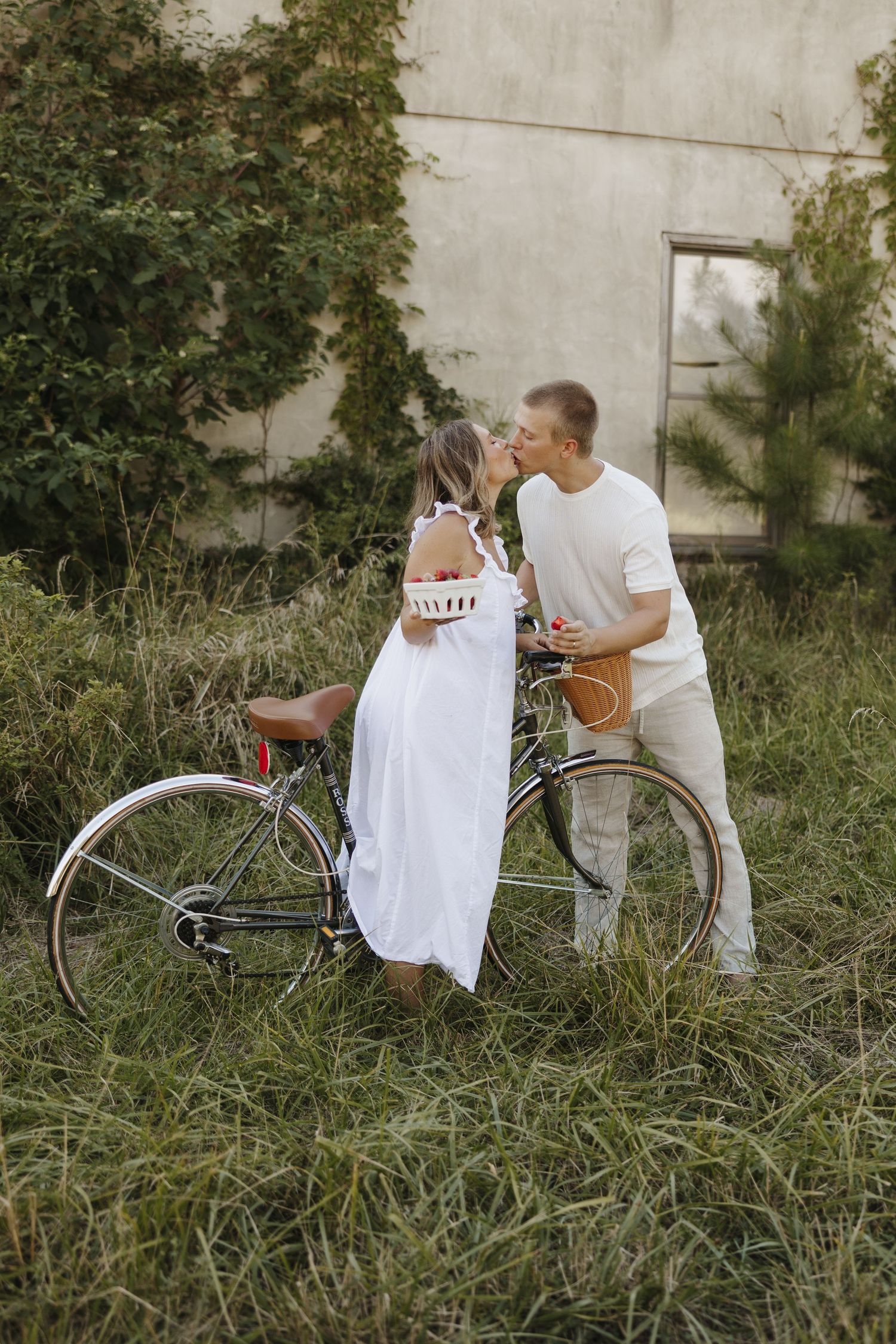 A romantic outdoor photoshoot with a couple sitting in tall grass next to a vintage bicycle against a white wall.