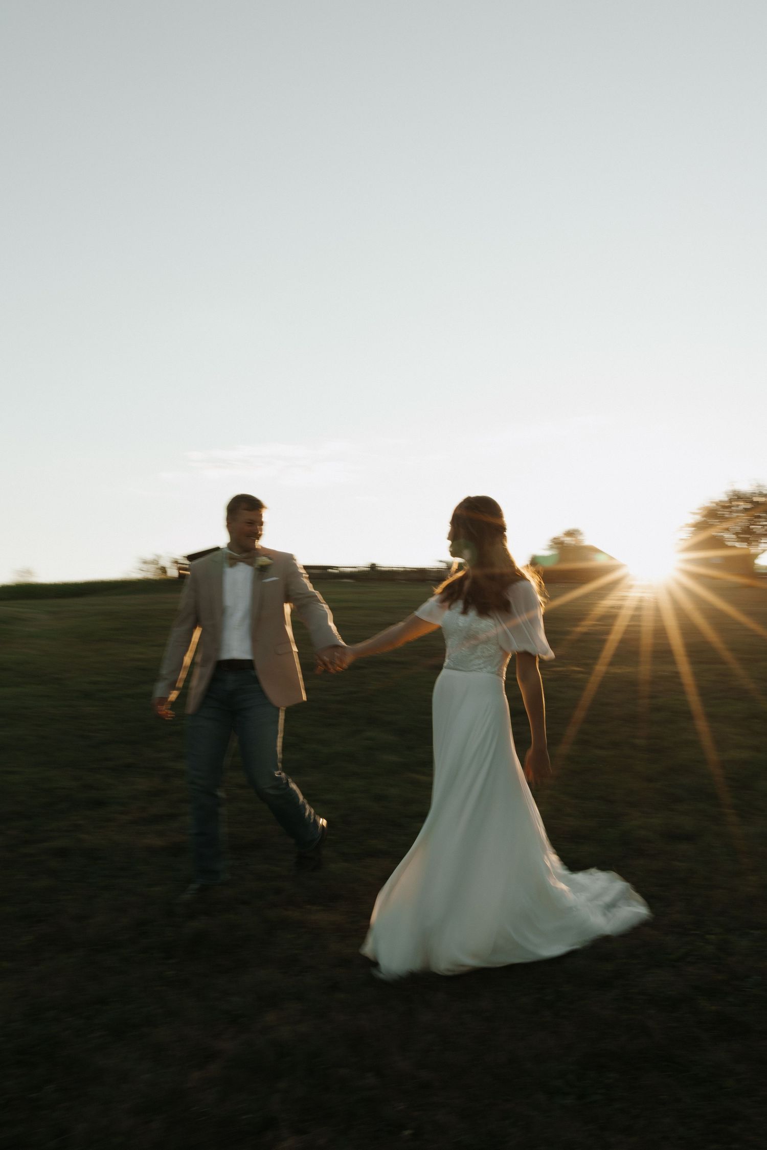 A couple in wedding attire dances together in a field at sunset creating a romantic silhouette against the sky.