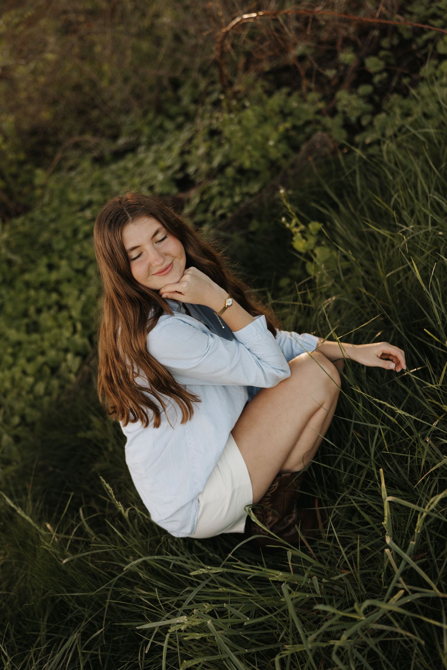 A woman in a white sweater sits in tall grass during a moody outdoor portrait session at dusk.