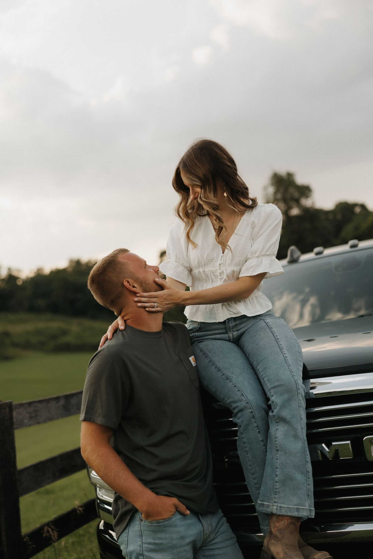 A romantic couple shares an intimate moment leaning against a black truck in a rural setting at dusk.