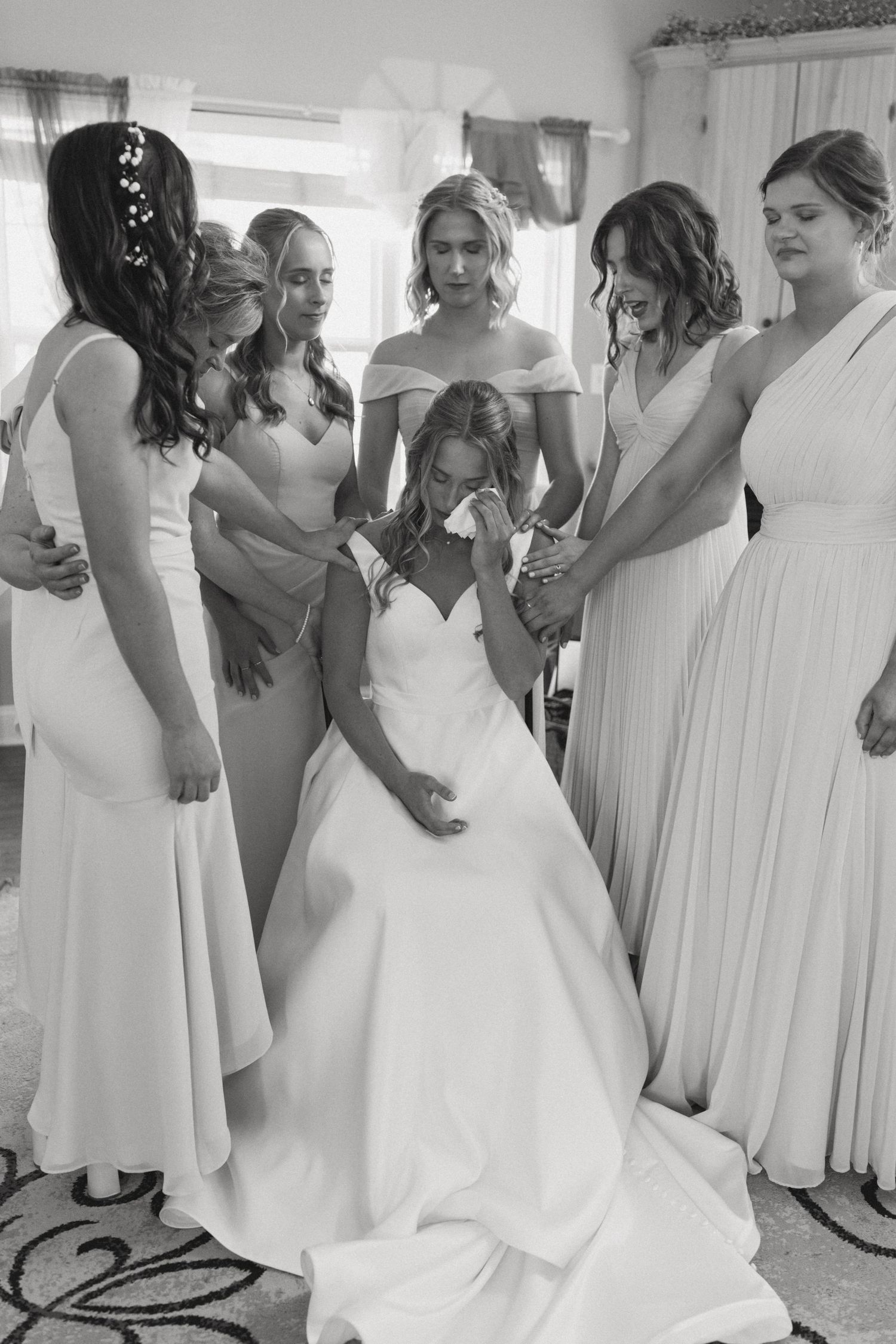 Bridesmaids in light colored dresses gather around the bride in her white gown during wedding preparations.