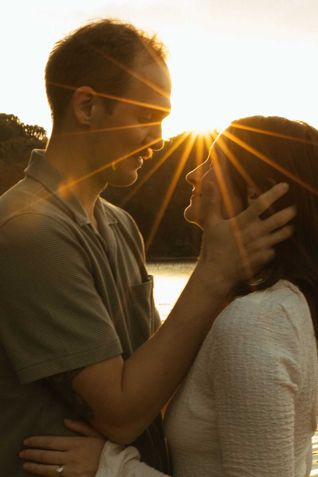 A romantic silhouette of a couple embracing at sunset by a lake with golden sunbeams shining through.
