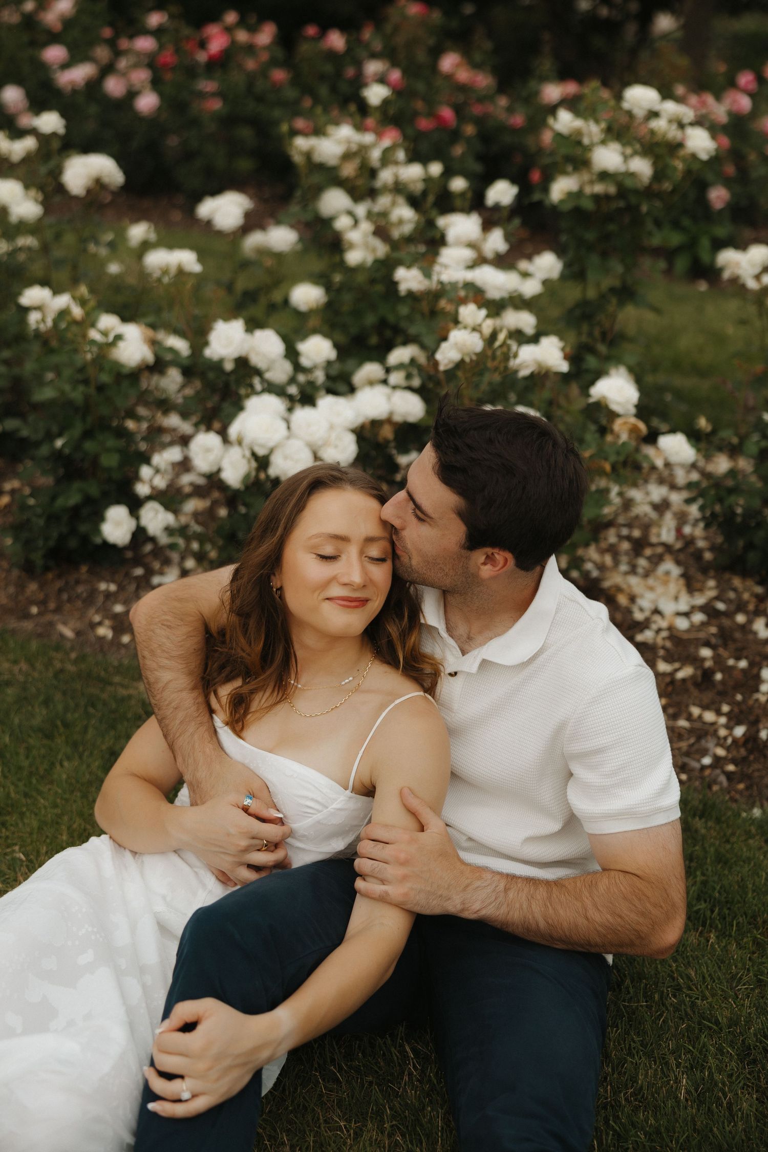 Romantic couple embraces on grass with blooming white and pink roses in garden background.