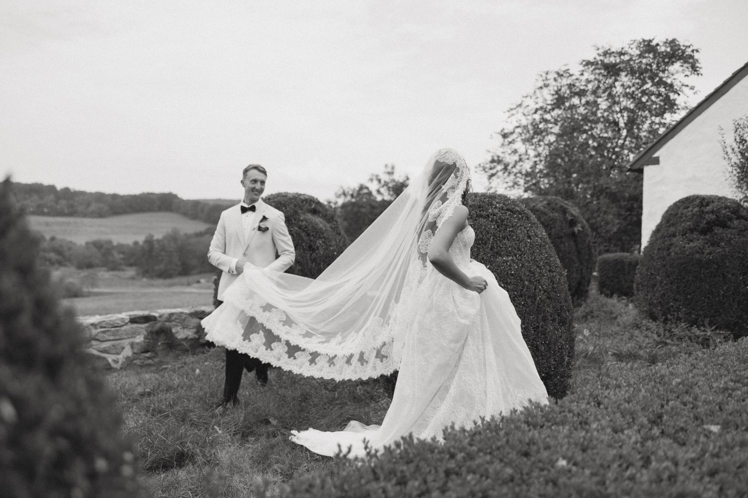 A wedding couple poses in a scenic countryside setting with a flowing veil creating an ethereal effect.
