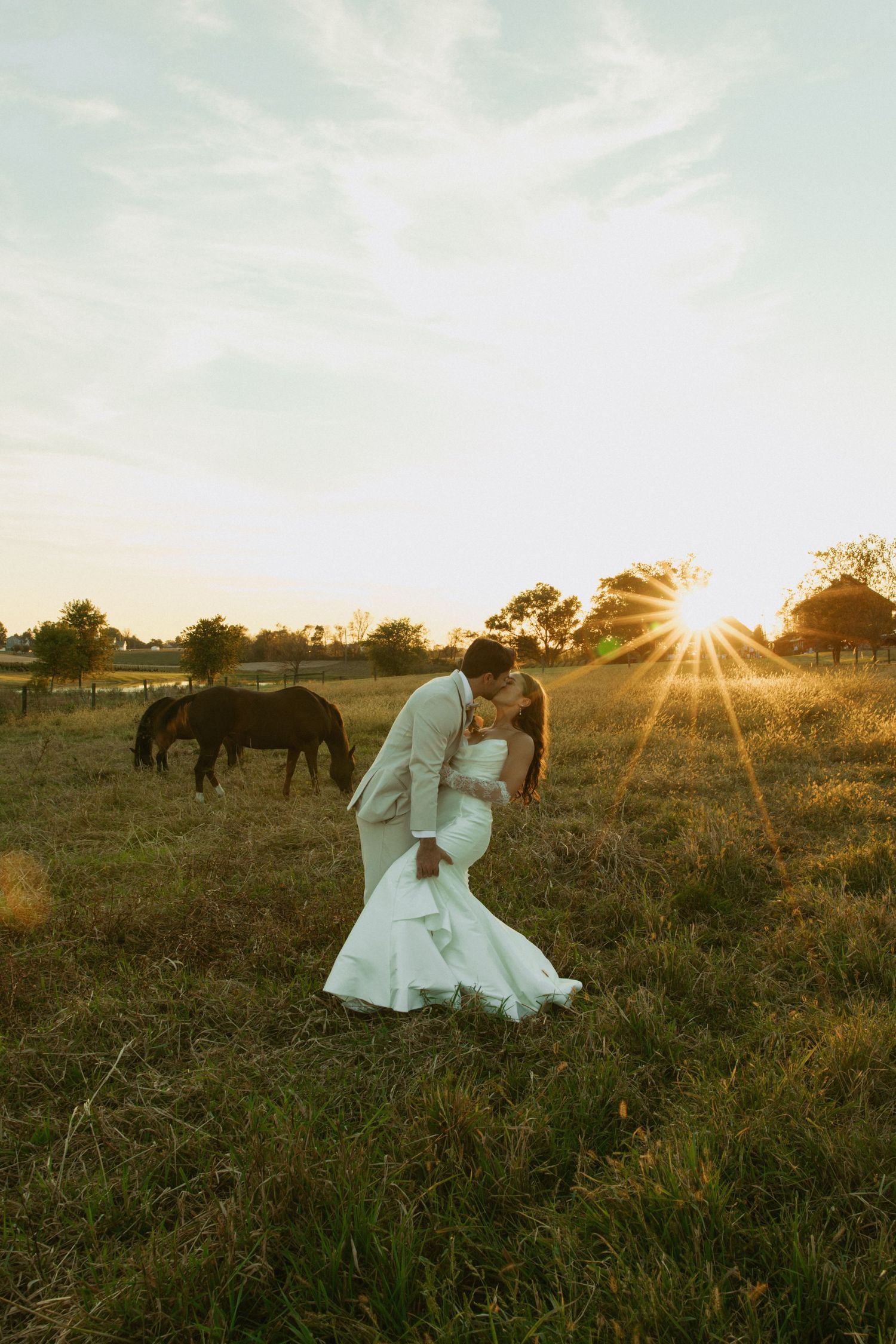 Bride and groom share a romantic dip kiss at golden sunset on a rustic farm with horses grazing in the background.