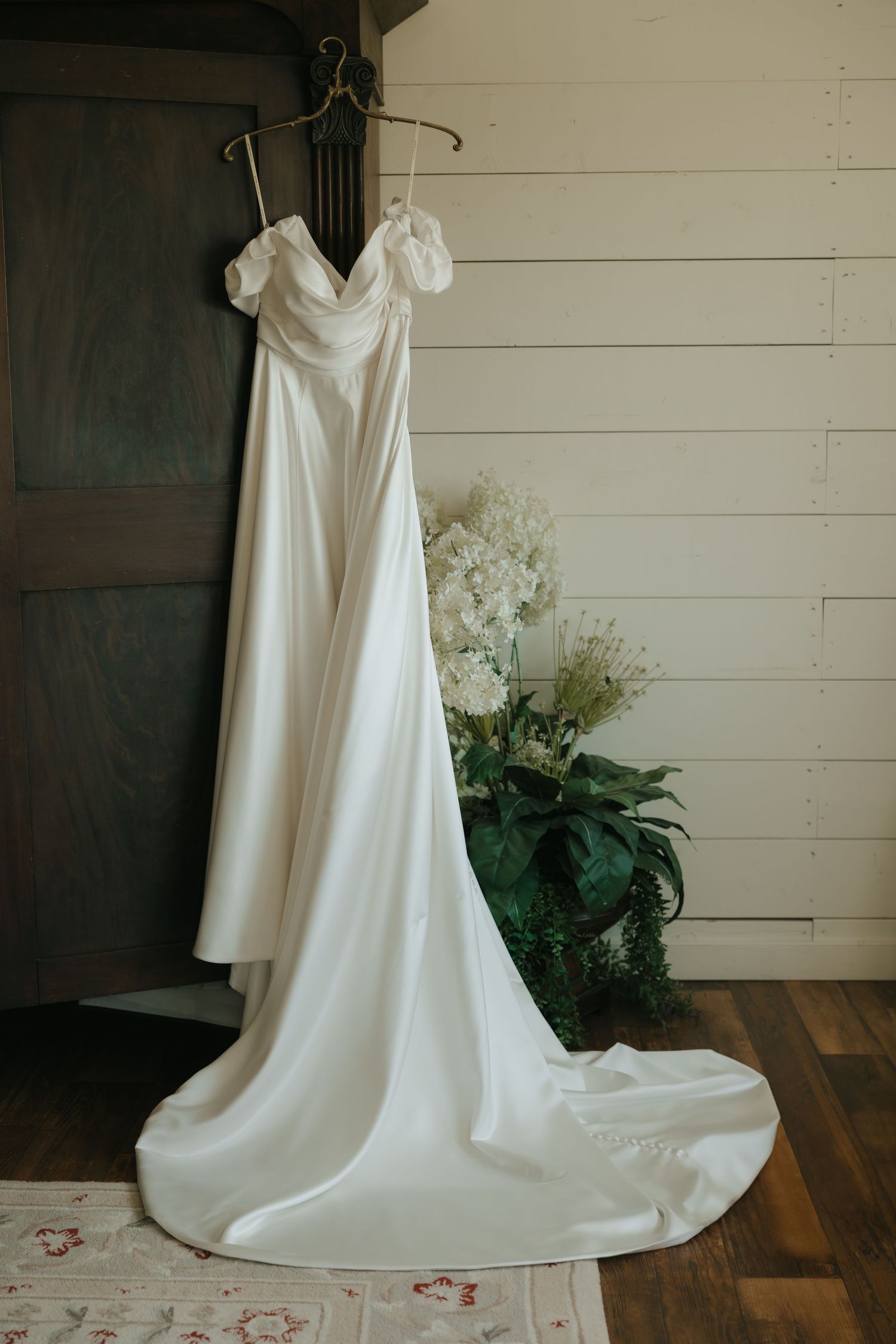 An elegant white wedding dress with off-shoulder design hangs on display next to potted flowers against a white shiplap wall.