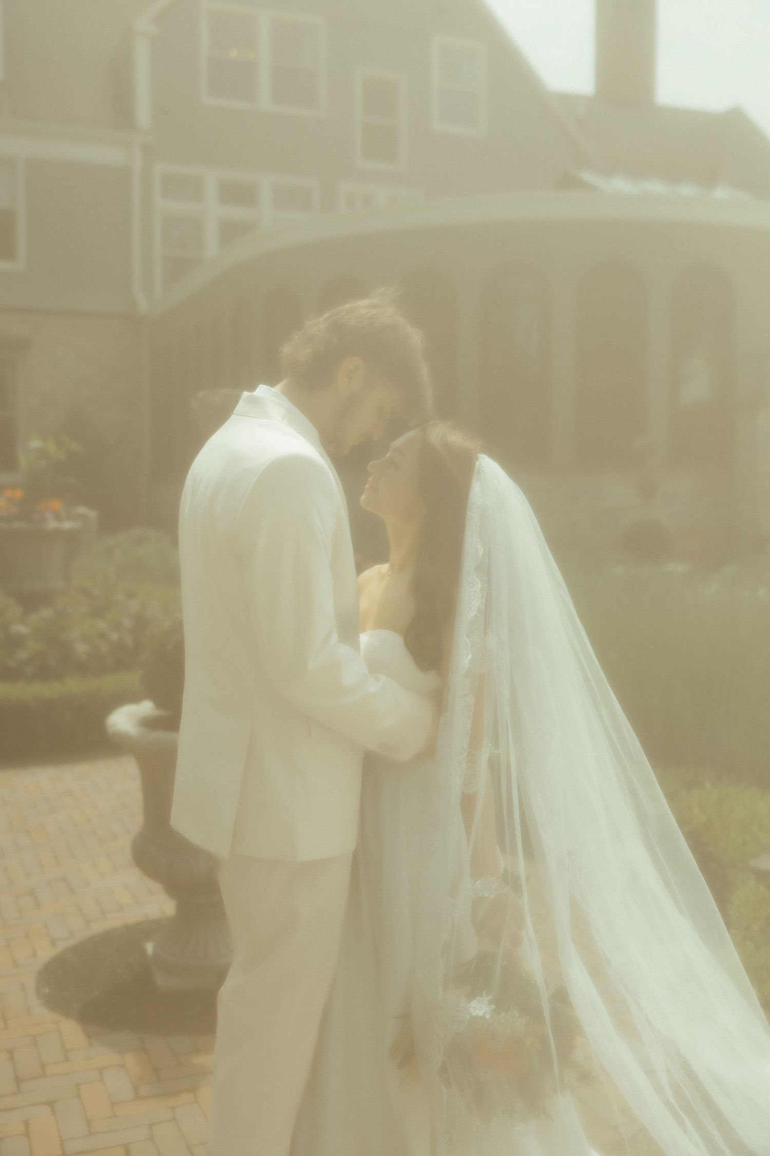 A romantic wedding couple embraces in a soft, dreamy outdoor setting with a flowing white veil catching the sunlight.