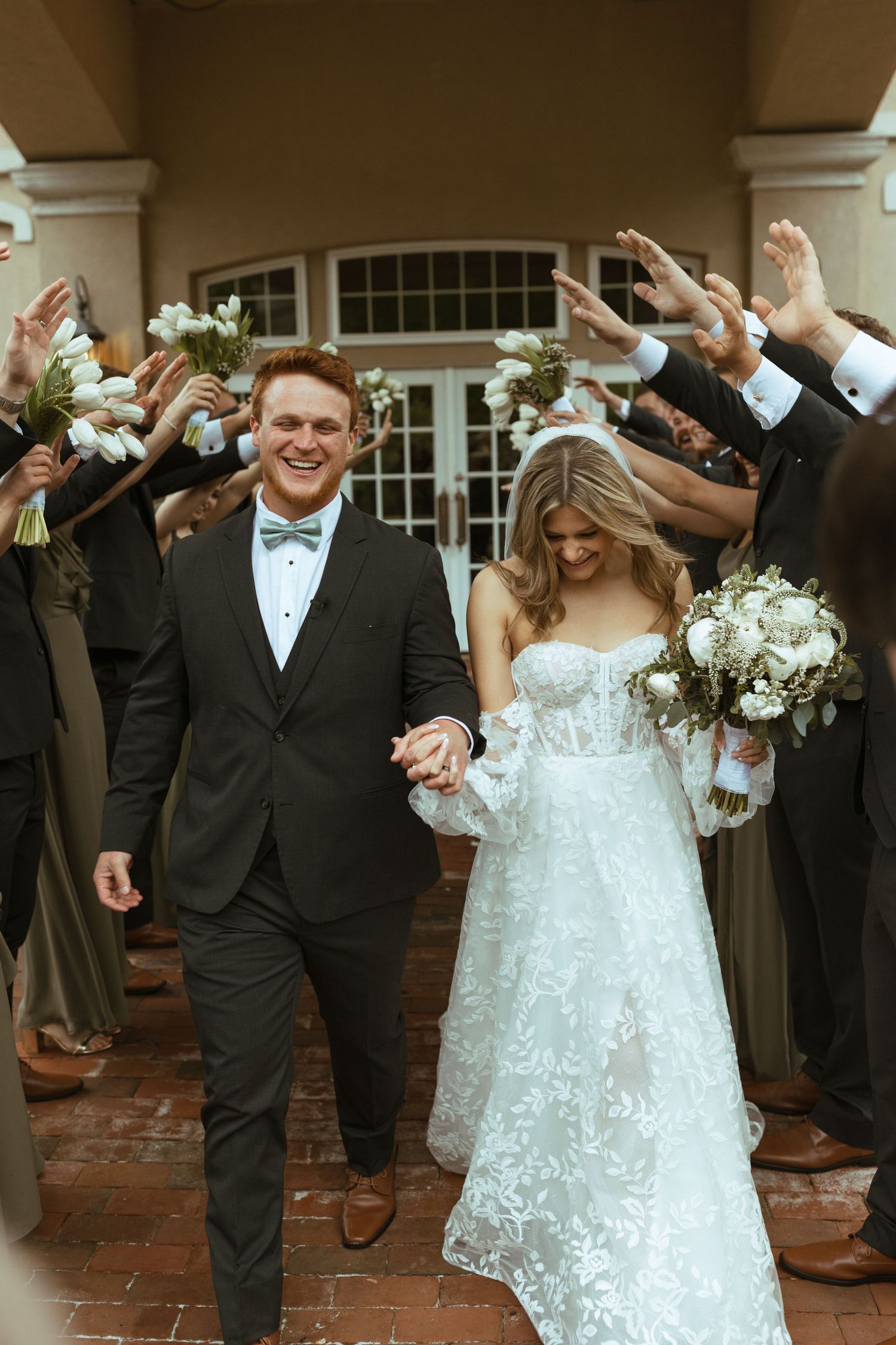 Wedding couple exits ceremony venue while guests throw confetti and cheer along a brick walkway.
