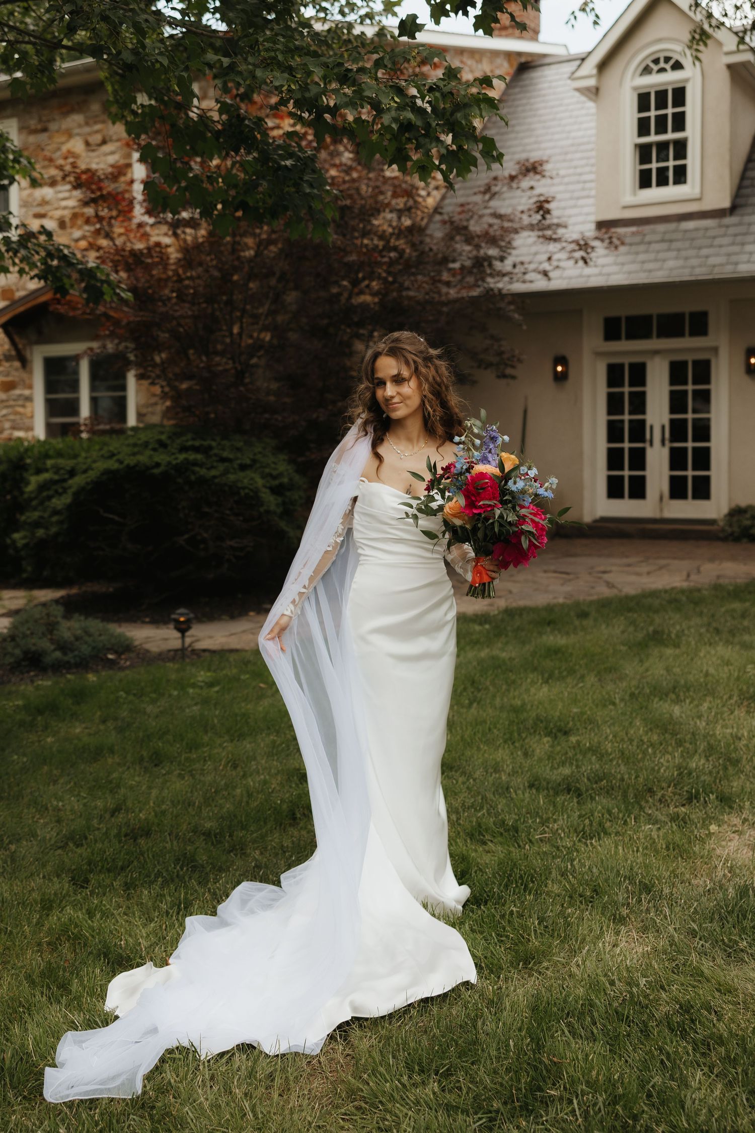Bride in flowing white dress walking on grass in front of a white house.