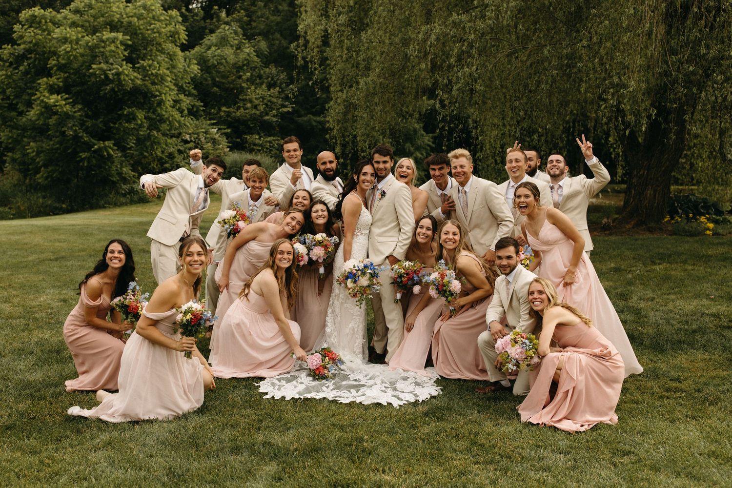 Wedding party in blush pink dresses poses together on grassy lawn during outdoor summer celebration.