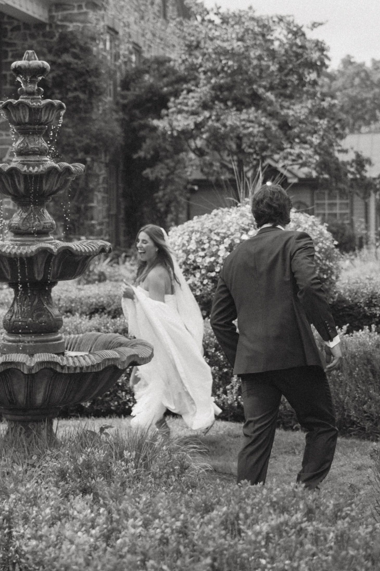 Black and white wedding photo showing couple walking through garden with ornate fountain.
