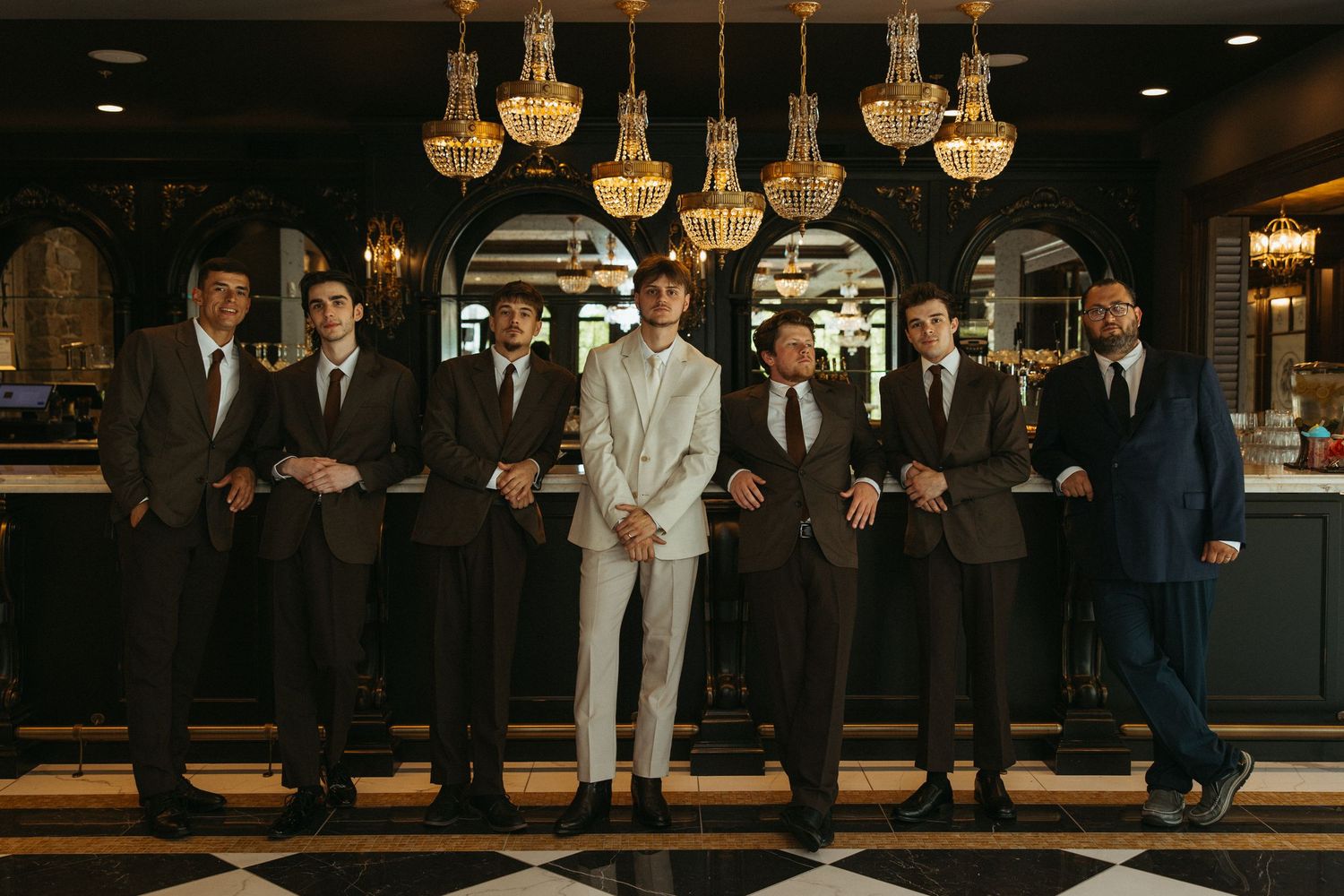 A group of formally dressed people stand together in an elegant room with ornate chandeliers overhead.
