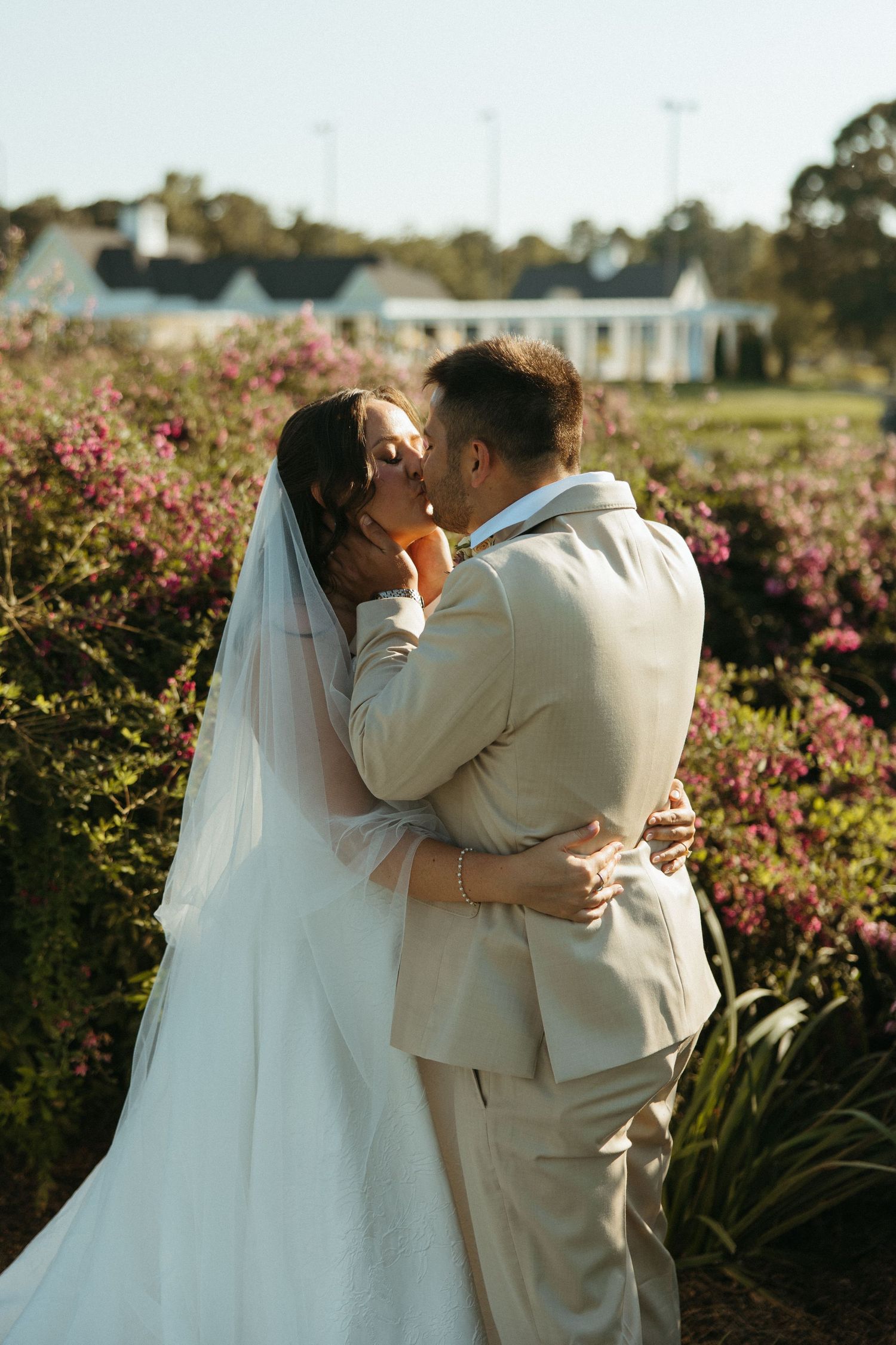 Newlywed couple shares romantic kisses in a garden with blooming pink flowers during golden hour.