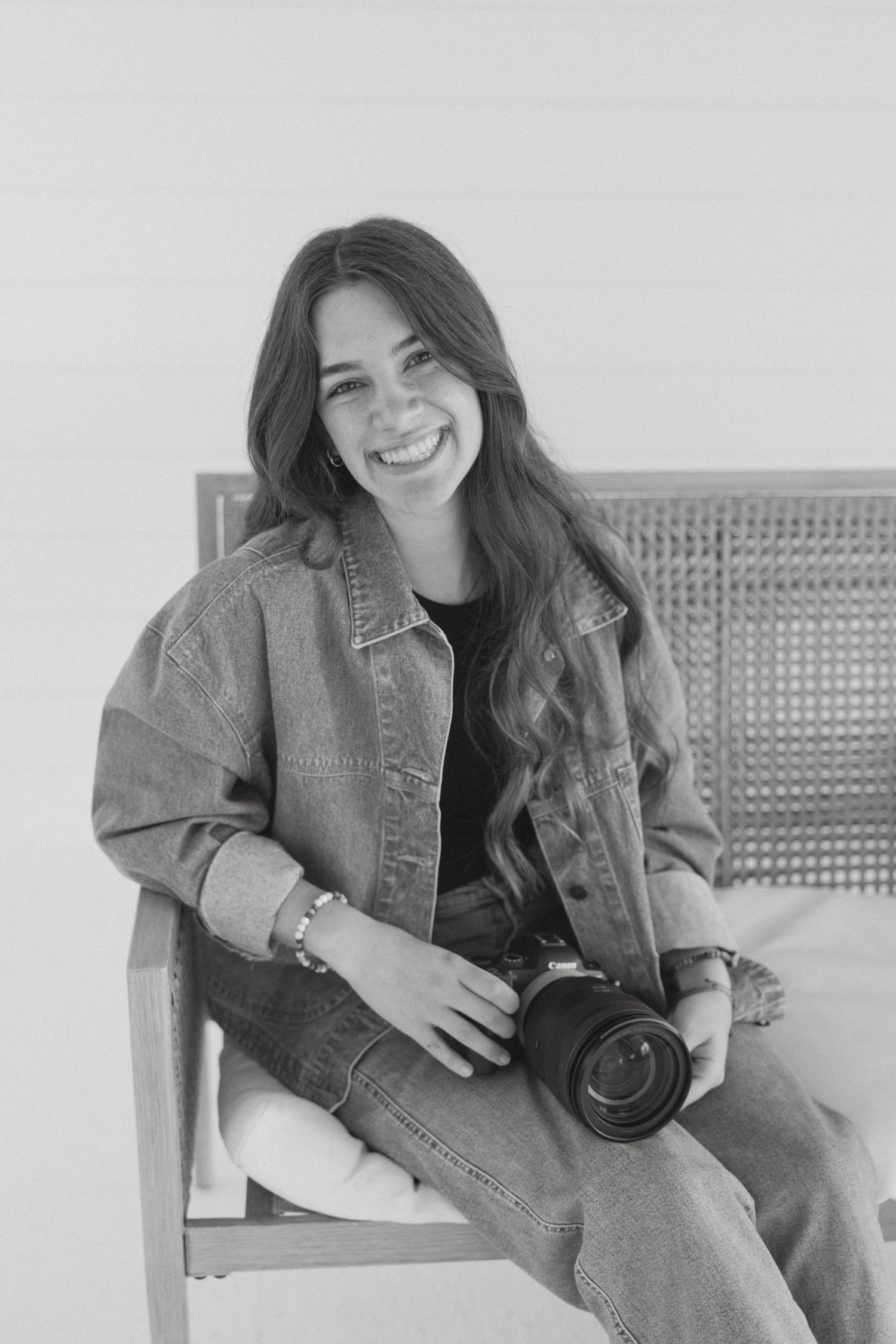 Black and white photo of someone in denim clothing sitting casually on a chair while holding a camera.